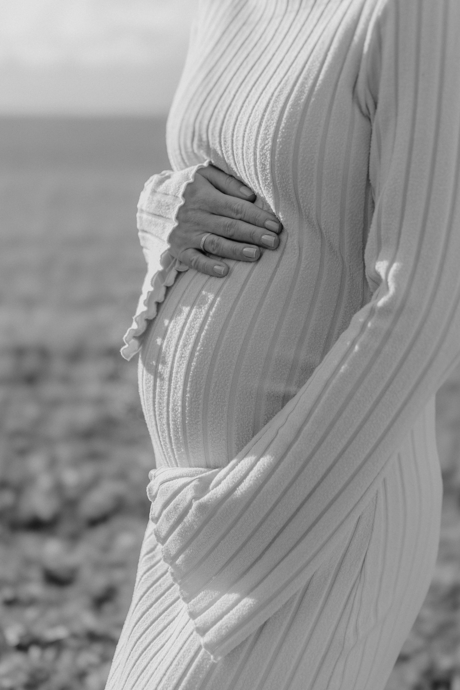 Black and white photo of a pregnant woman in a ribbed dress cradling her baby bump by the ocean.