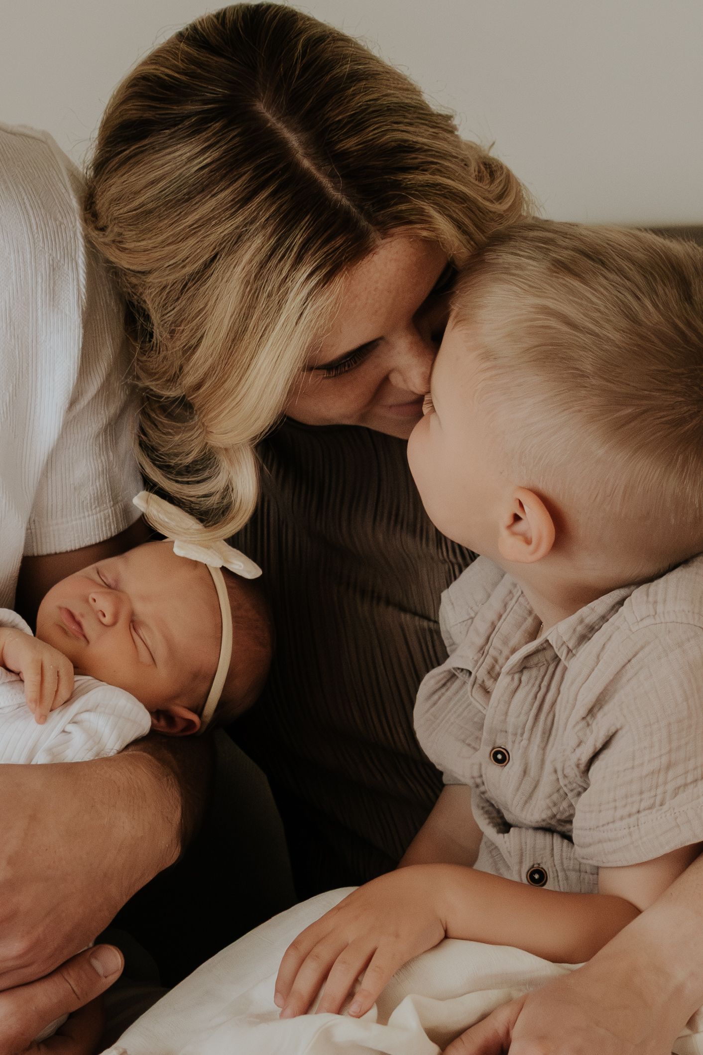 A mother tenderly kisses her toddler son while cradling her sleeping newborn baby in a warm family portrait.
