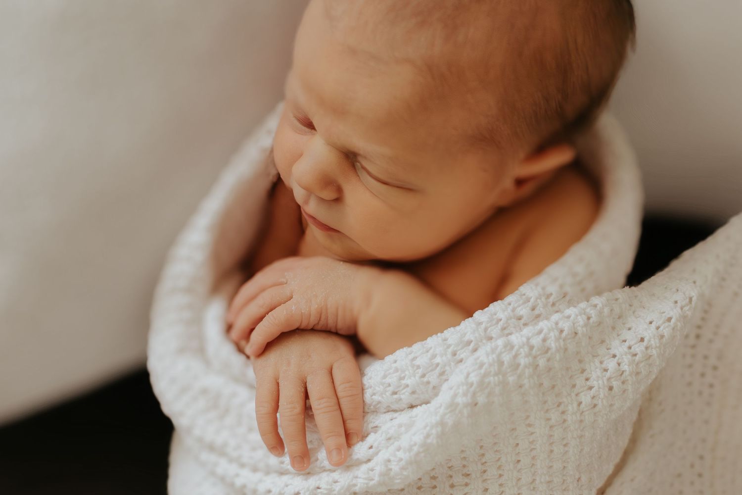 Peaceful newborn baby sleeping wrapped in a soft white knit blanket, tiny fingers visible in a close-up portrait.