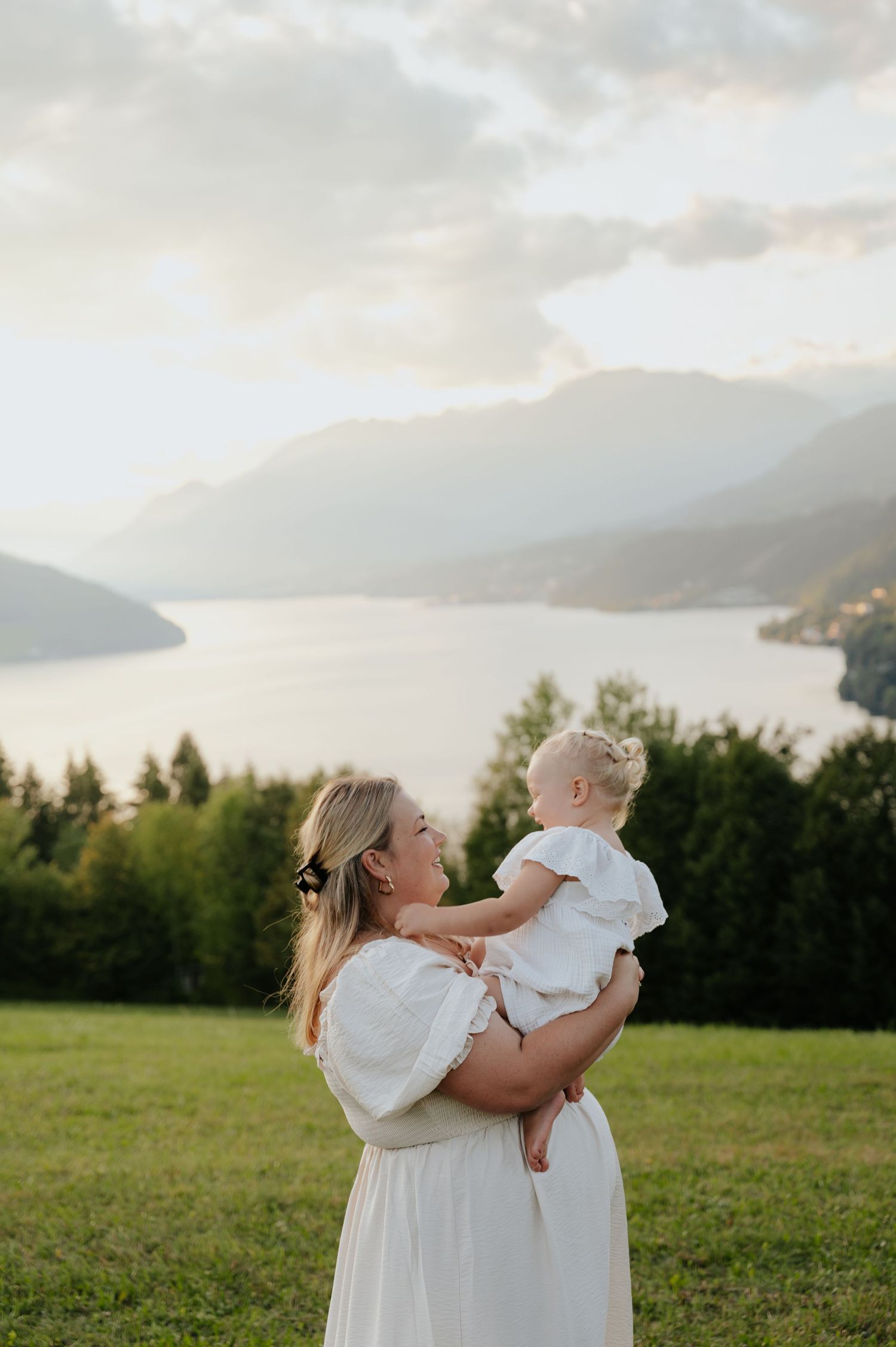 Mother in white dress holding baby girl overlooking scenic mountain lake at golden hour.