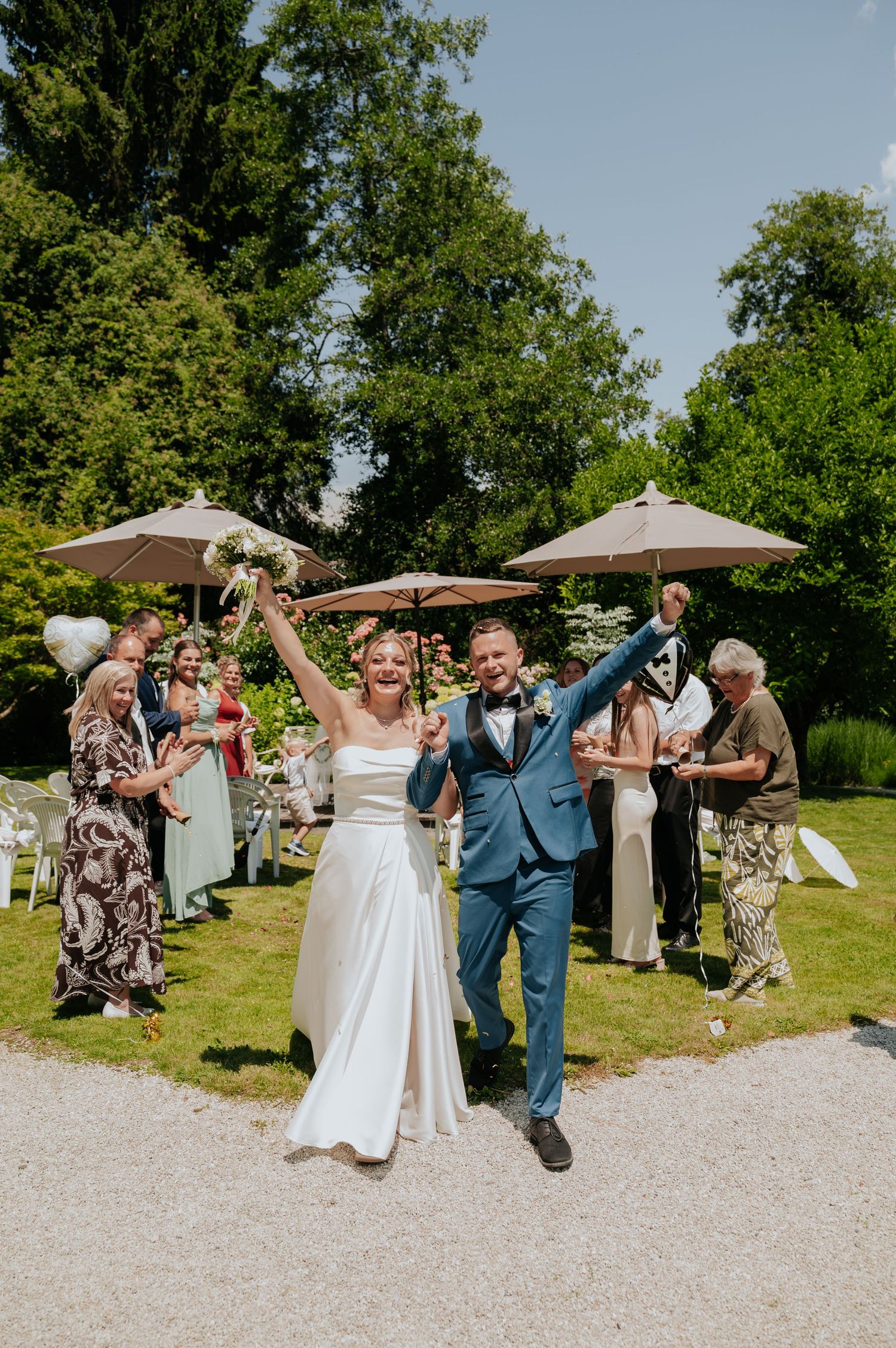 Joyful newlyweds celebrate their outdoor wedding ceremony, raising arms as guests cheer in a sunny garden.