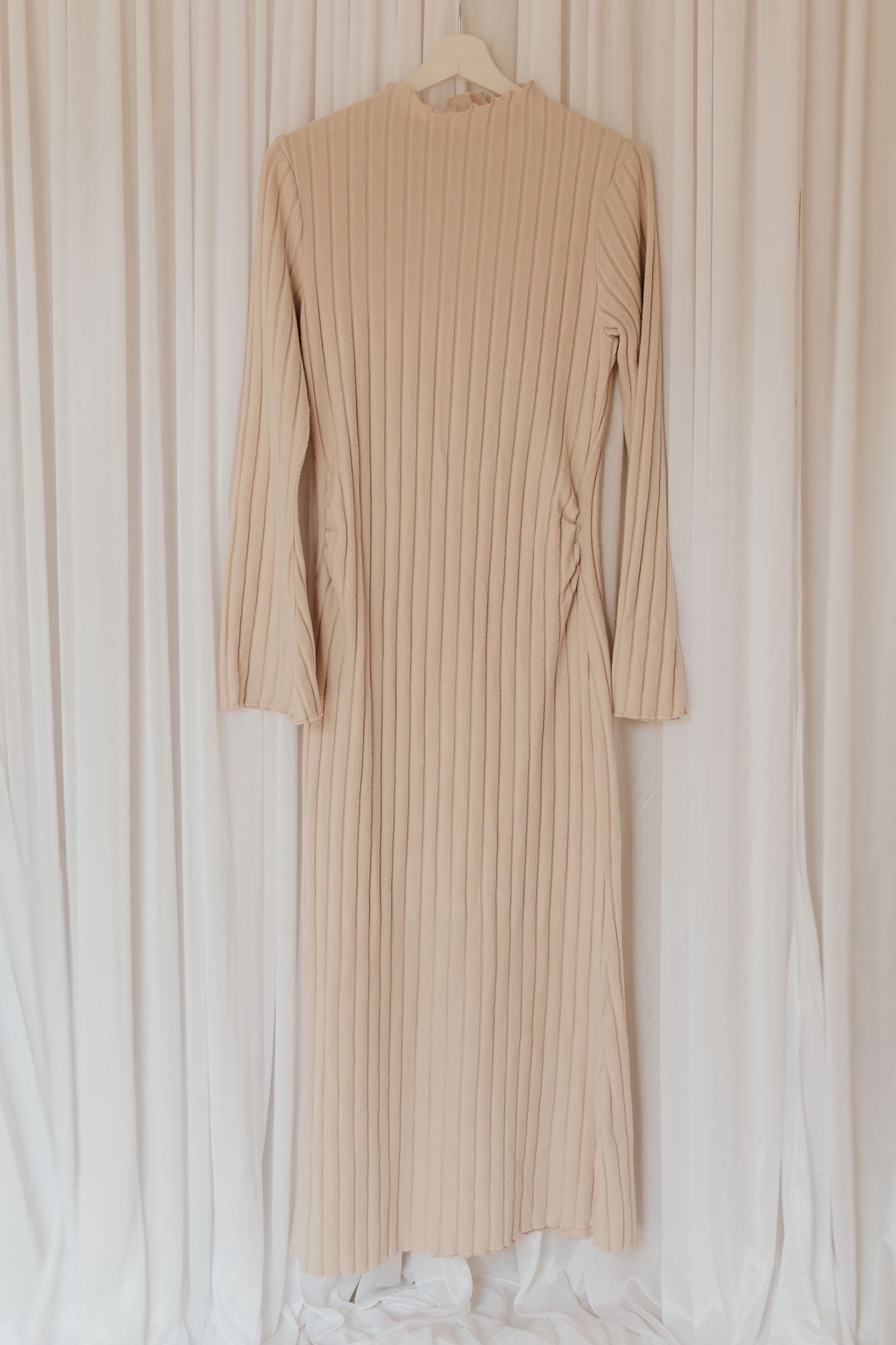 Beige geripptes Strickpulloverkleid, das vor weißem Vorhanghintergrund hängt.