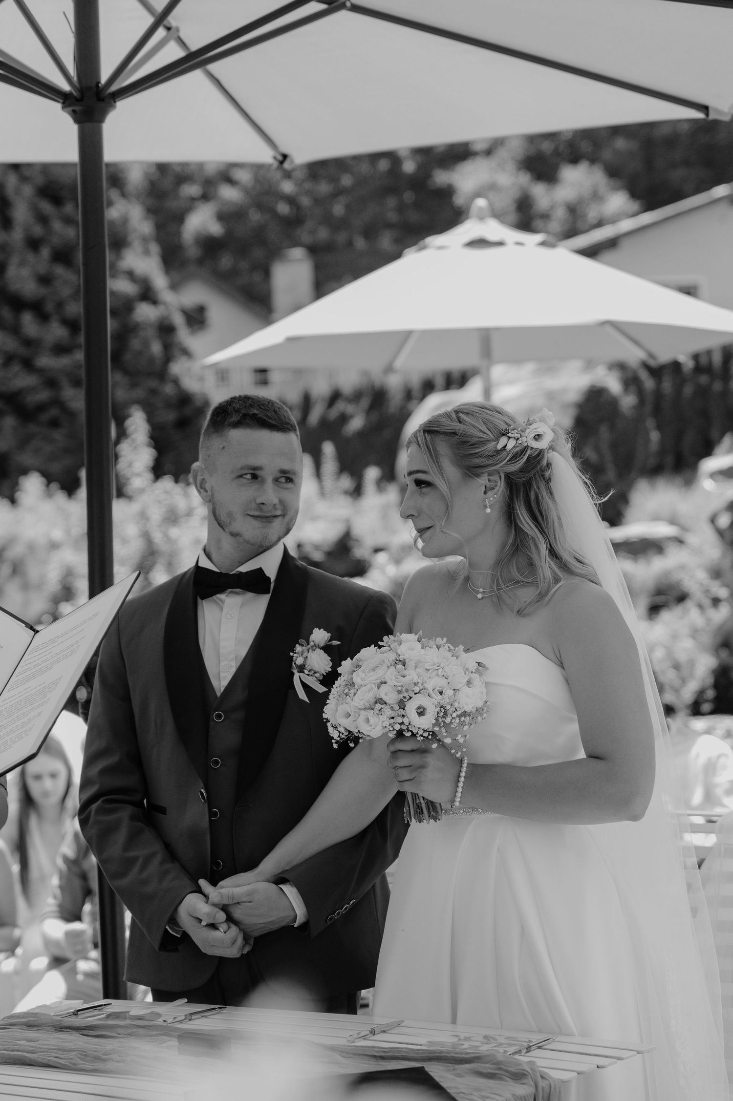 Black and white photo of a smiling bride holding a bouquet and groom in a tuxedo at an outdoor wedding ceremony.