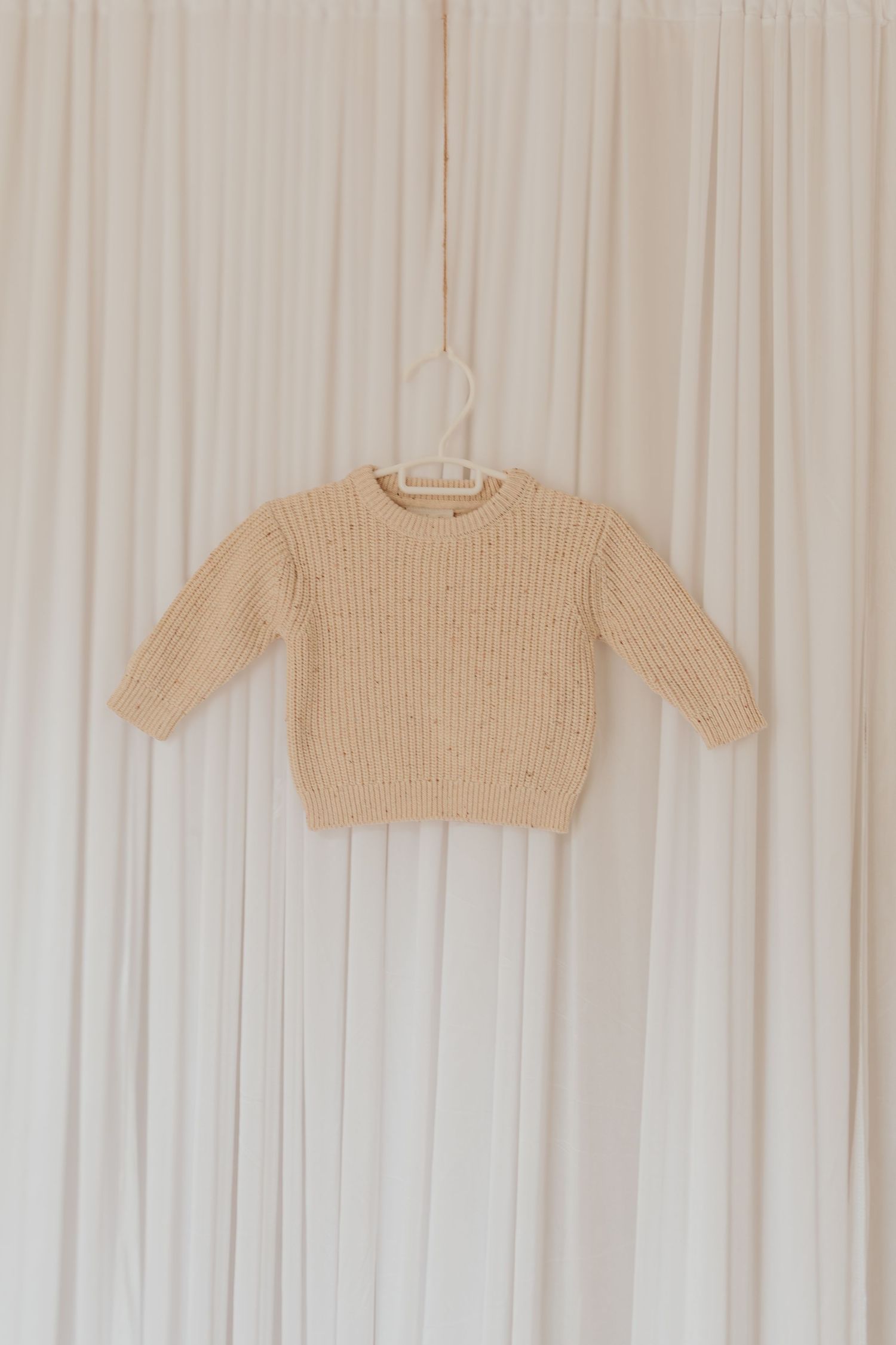 Beige gestrickte Babypullover, die in einer Reihe an weißen Kleiderbügeln vor einem weißen Vorhanghintergrund hängen.