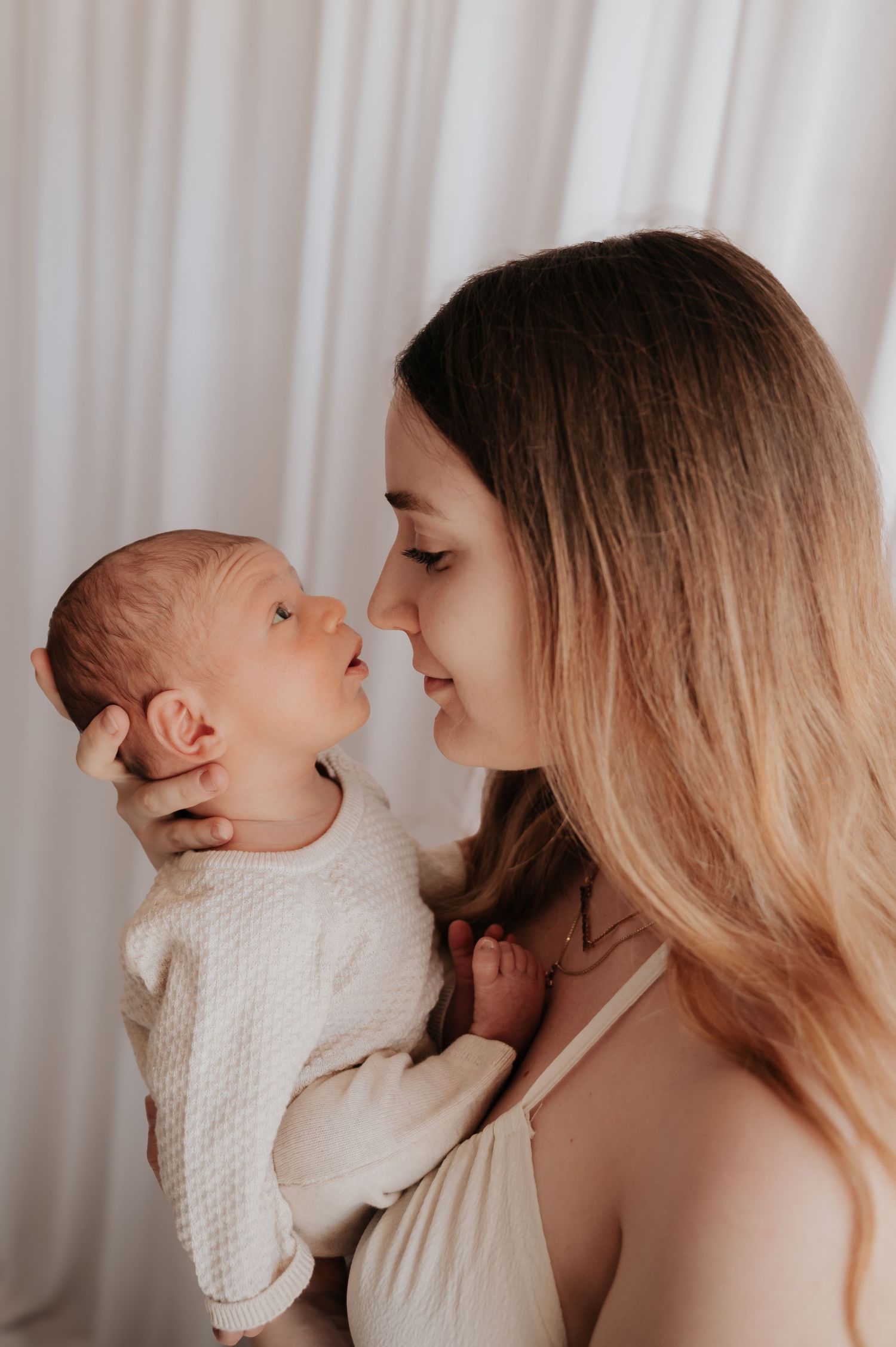 A young mother tenderly holds her newborn baby close, sharing a gentle nose-to-nose moment in soft white light.