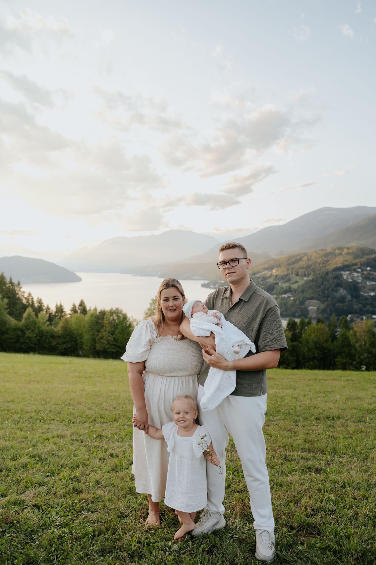 Family of four posing on green hillside with mountain lake backdrop at sunset, parents holding newborn baby and toddler.