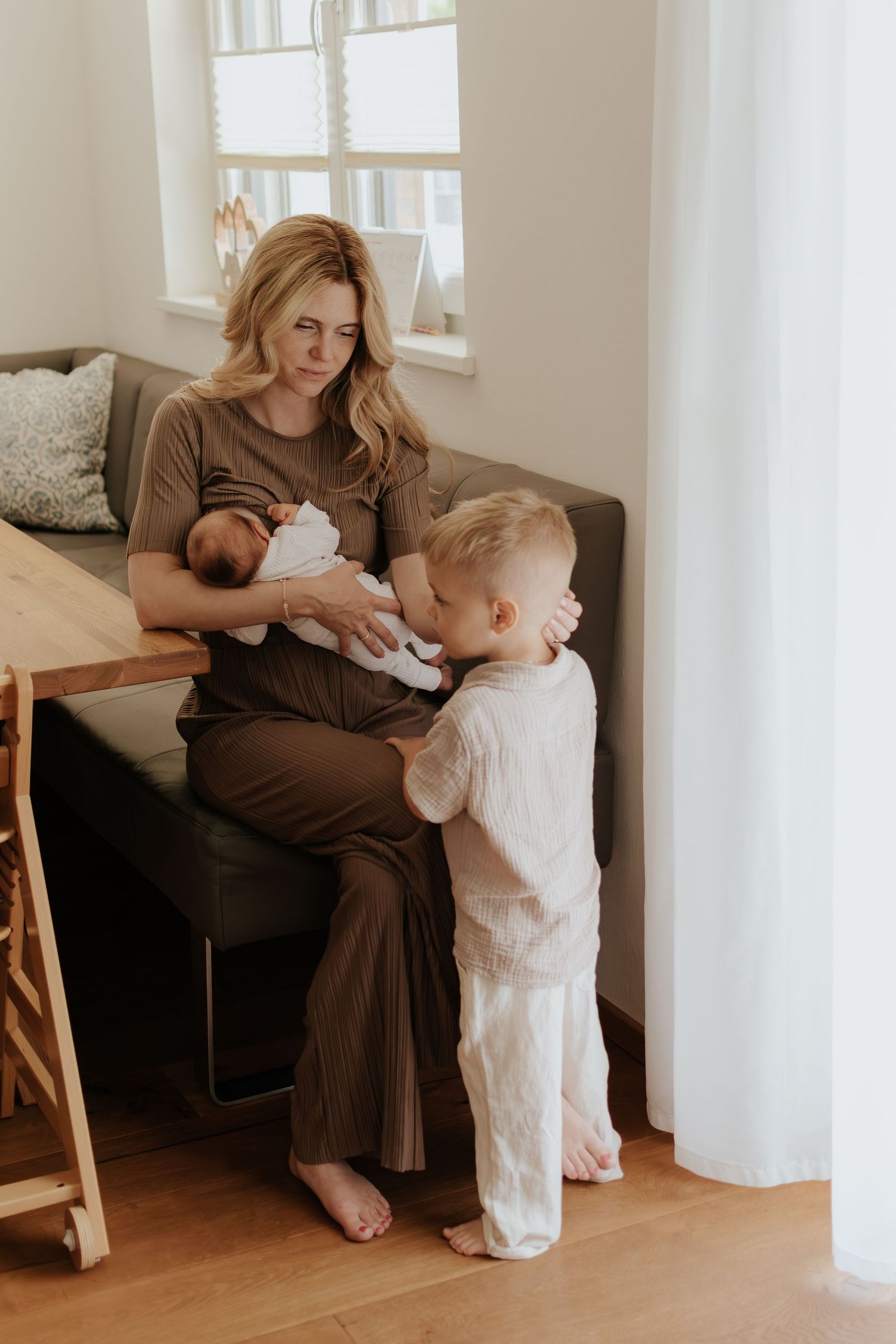 A mother sits on a gray sofa holding a newborn while a young toddler boy stands beside her in a bright home.