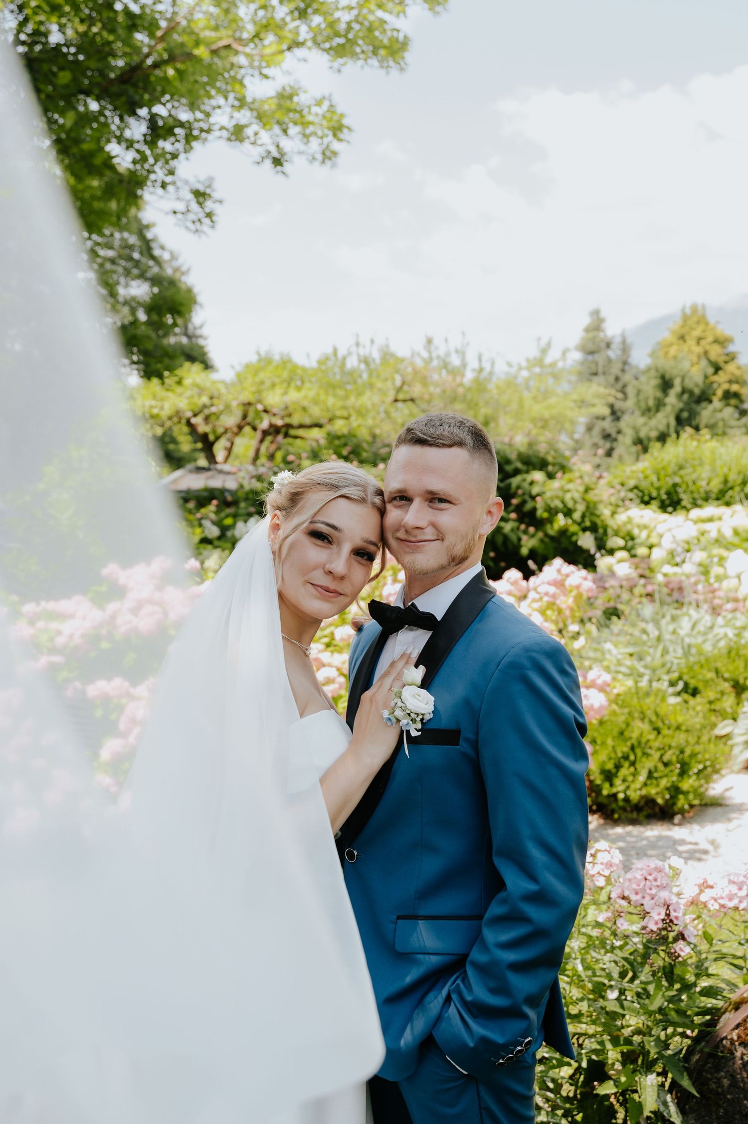 A bride and groom pose together in a blooming garden, she in a white veil and he in a blue tuxedo with bow tie.