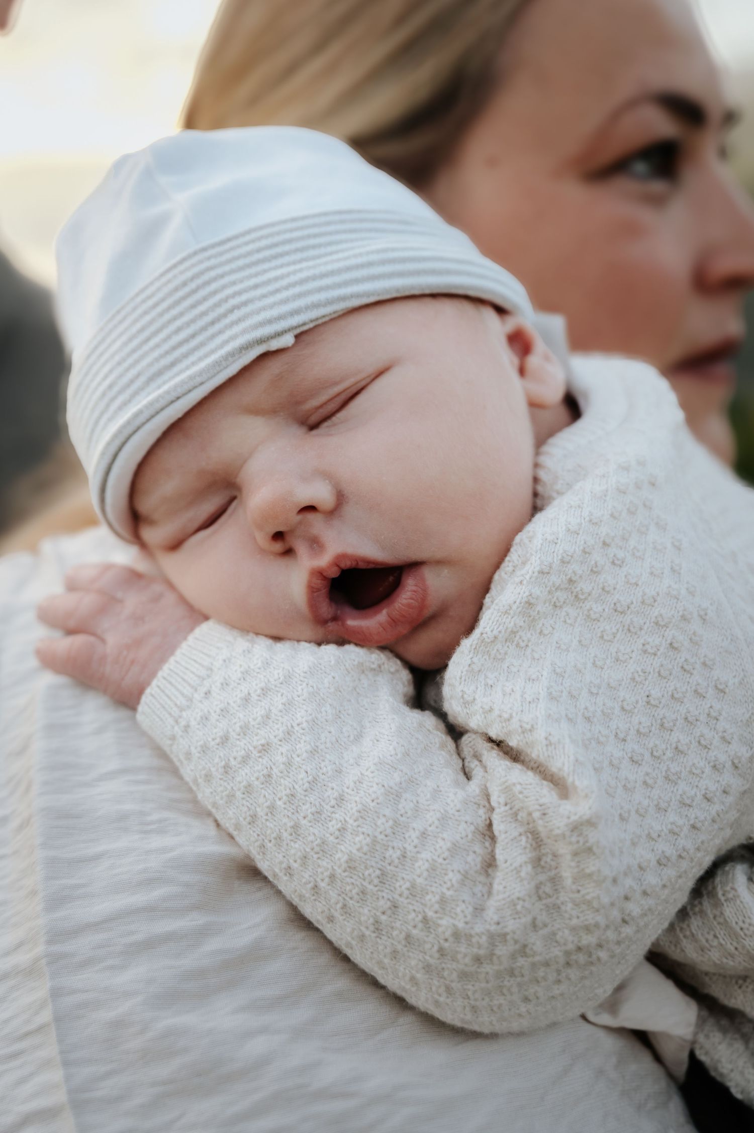 A newborn baby in a white knit outfit and striped hat yawns while being held on a mother's shoulder.