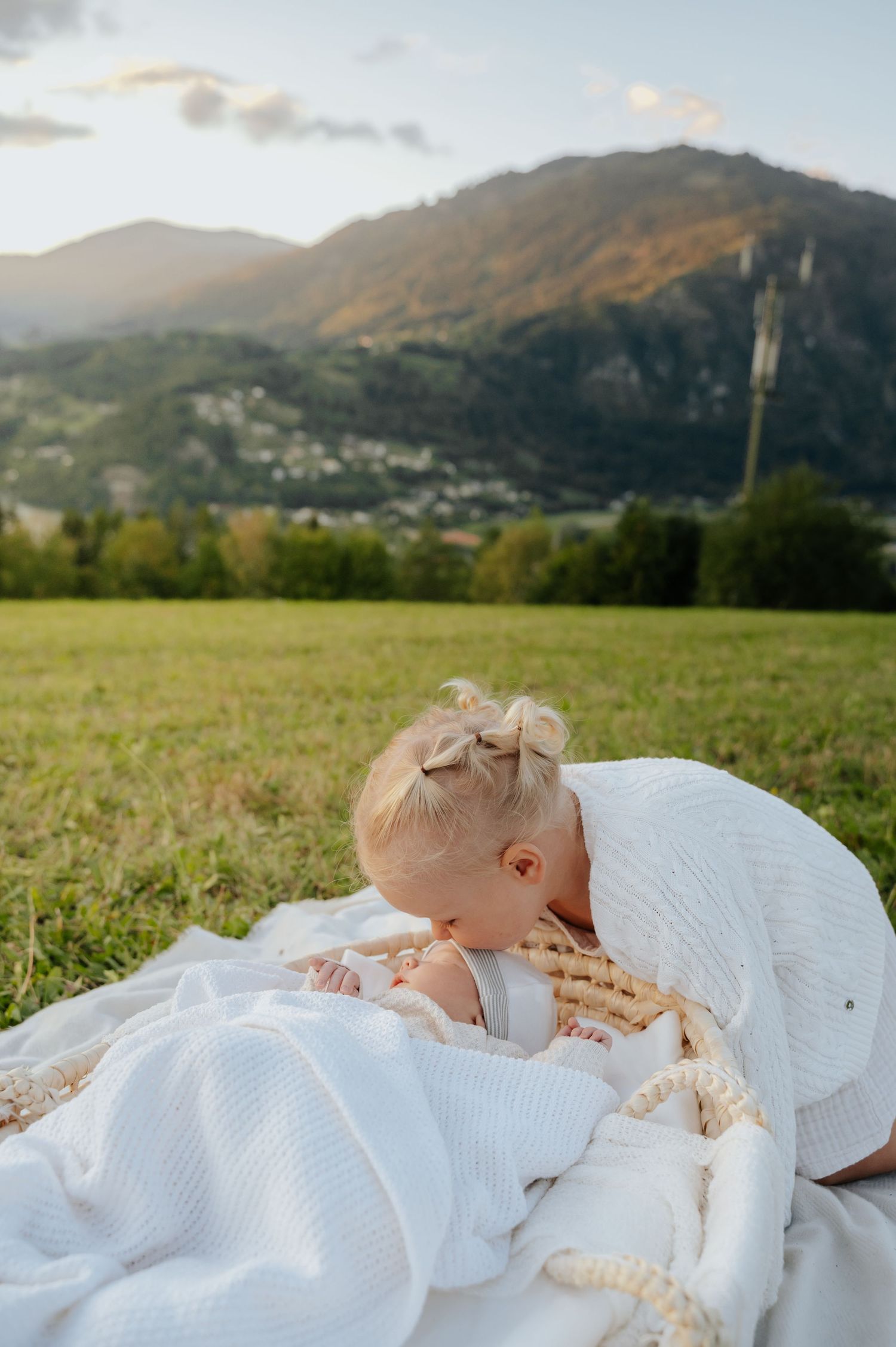 A toddler girl with bow hair ties bends to kiss a newborn baby on a white blanket in a scenic mountain meadow.