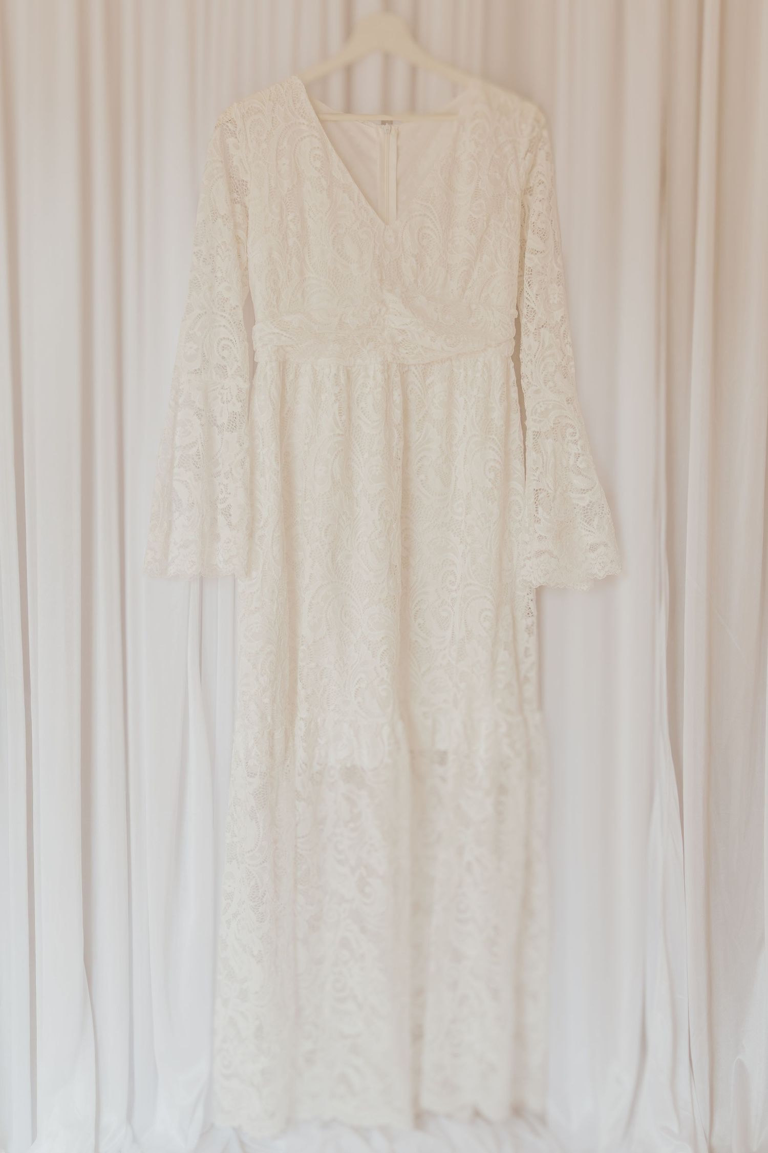 Ein weißes Spitzenkleid im Vintage-Stil mit langen Glockenärmeln und V-Ausschnitt hängt vor einem hellen Vorhanghintergrund.