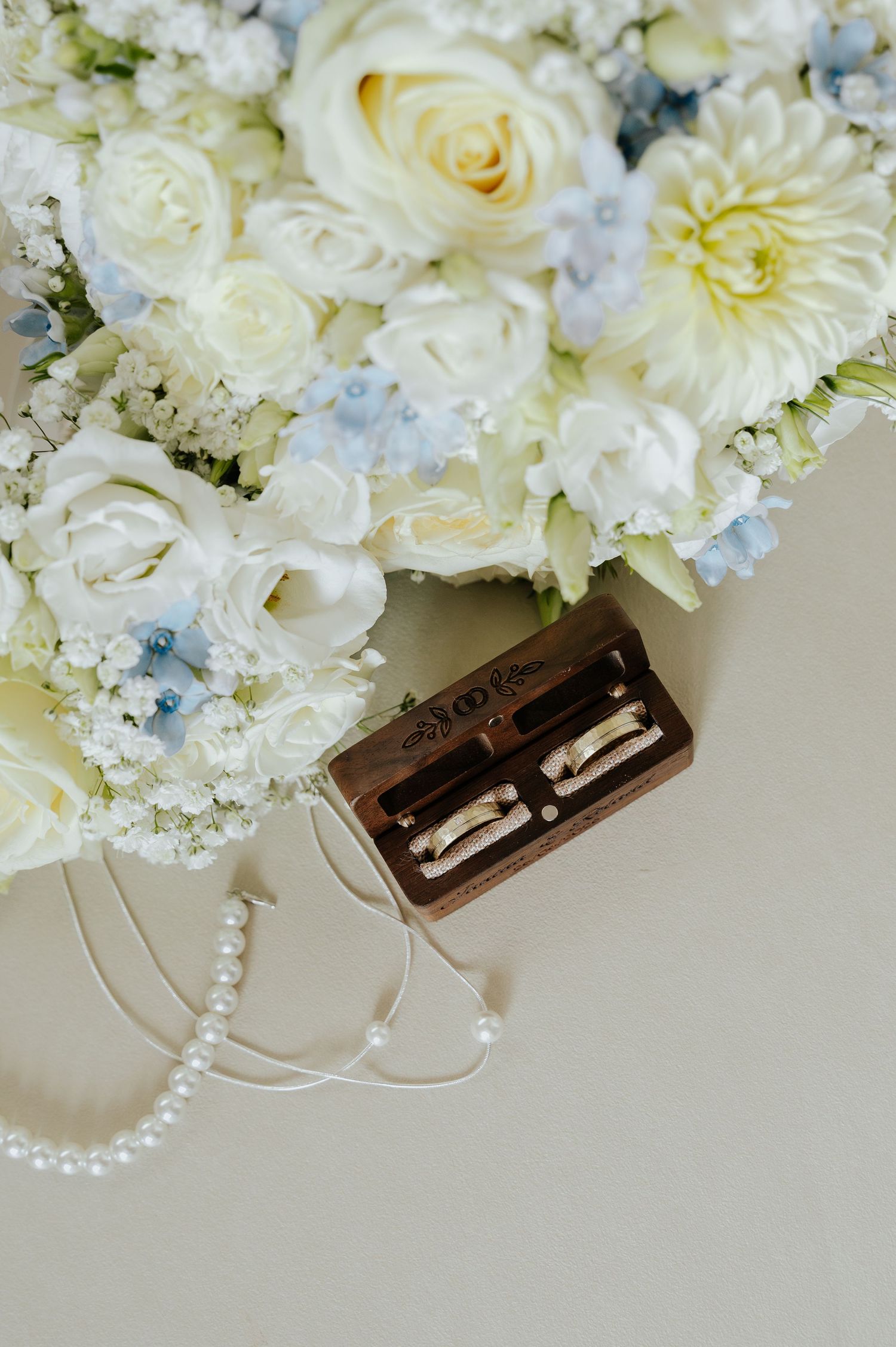 Wedding rings in wooden box beside white and blue floral bouquet with pearl necklace on neutral background.