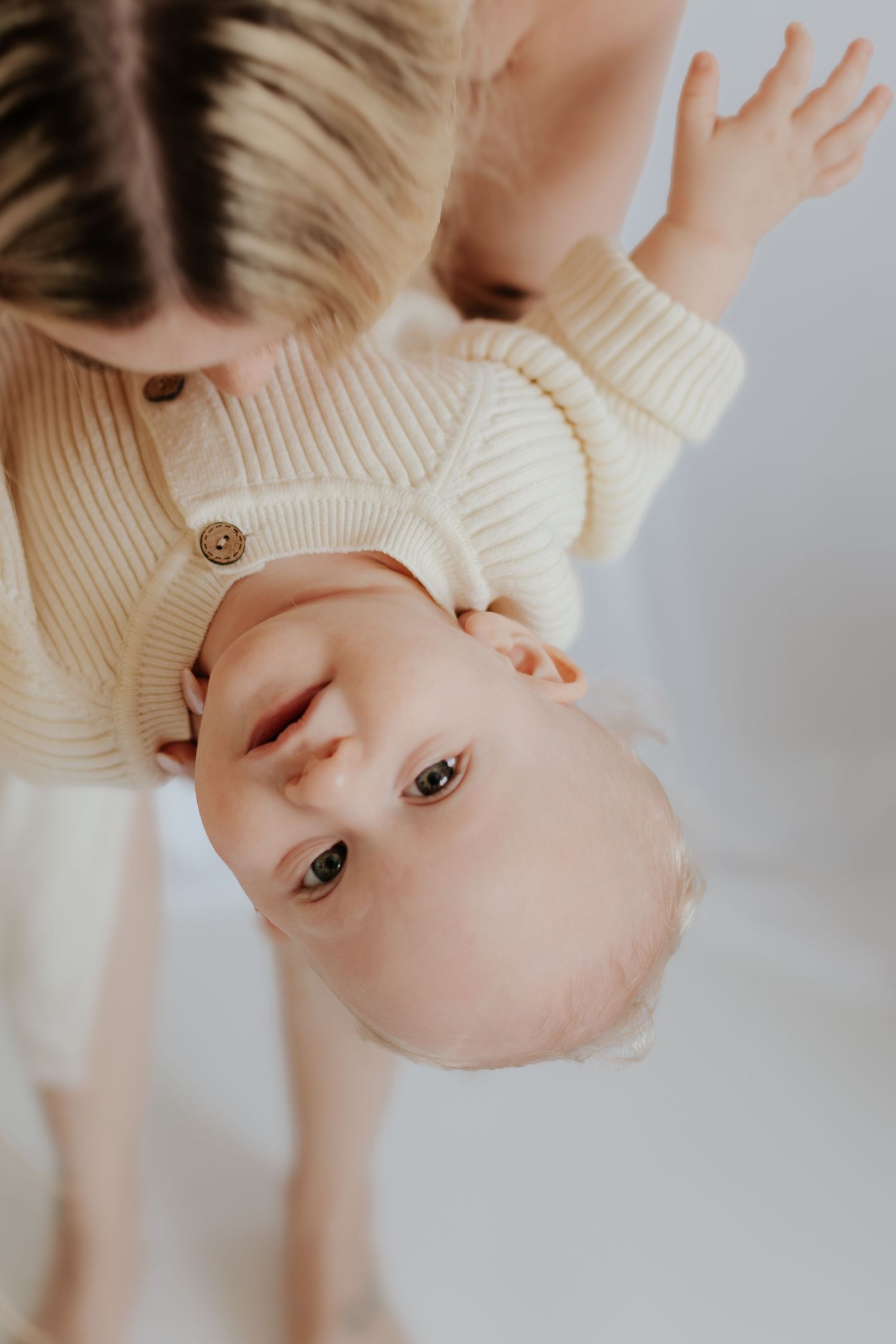 A baby in a cream knit romper is held upside down by a woman, looking up at the camera with curiosity.