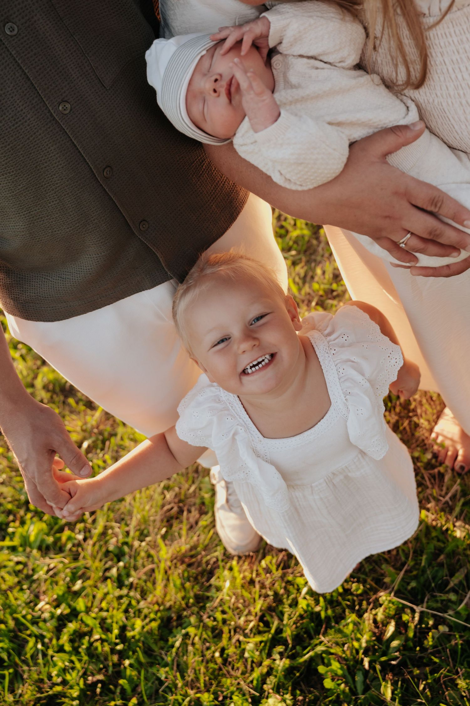 Smiling toddler girl in white dress holds parents hands outdoors while mother cradles newborn baby in golden sunlight.