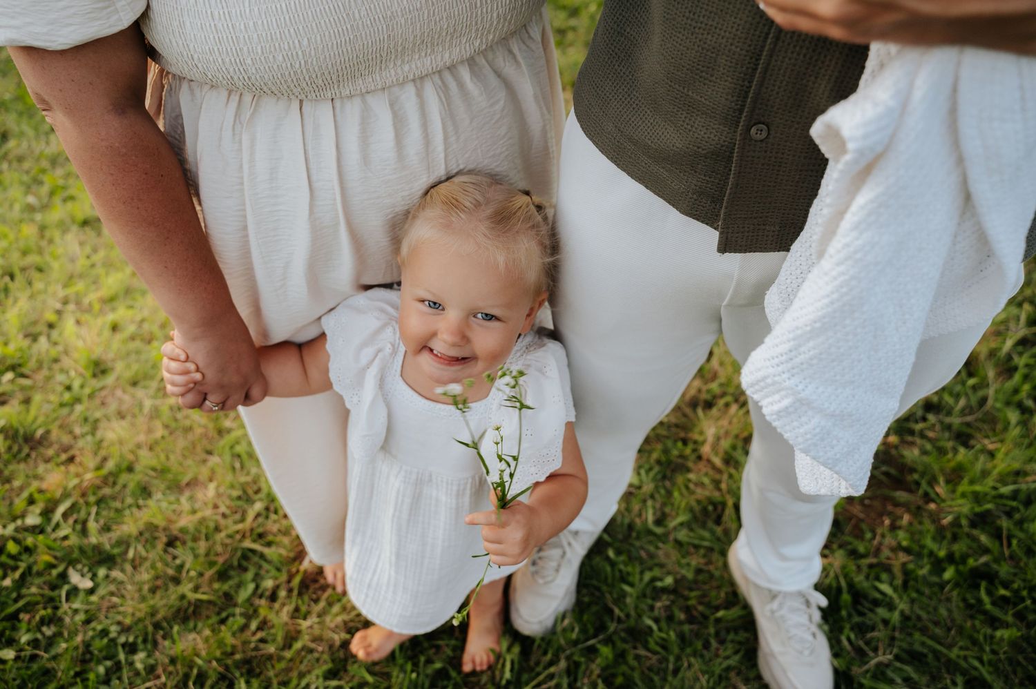 Baby in white dress holds wildflowers between parents on green grass during outdoor family photo session.