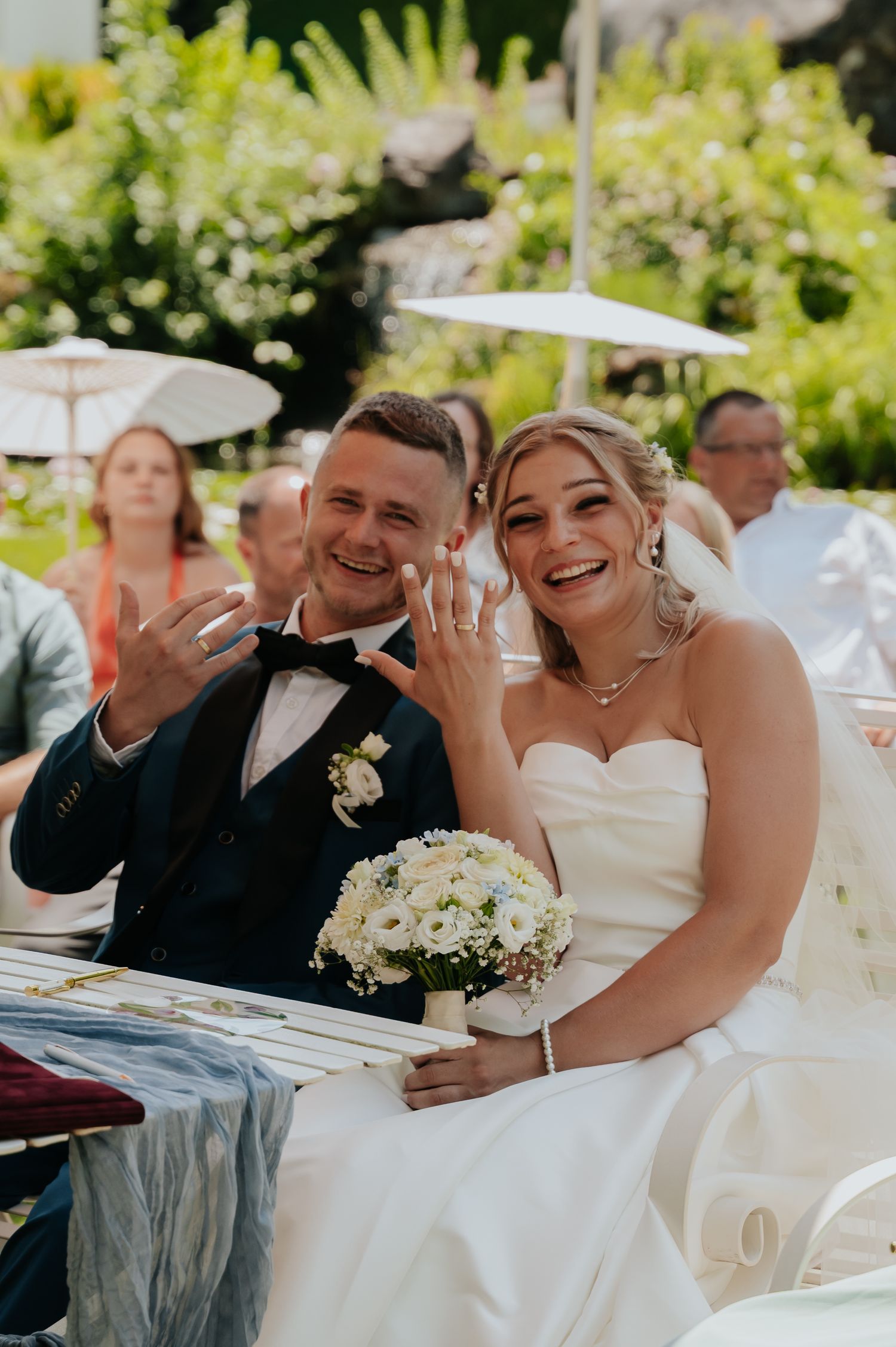Smiling bride and groom showing wedding rings at outdoor ceremony, bride holding white floral bouquet in strapless gown.