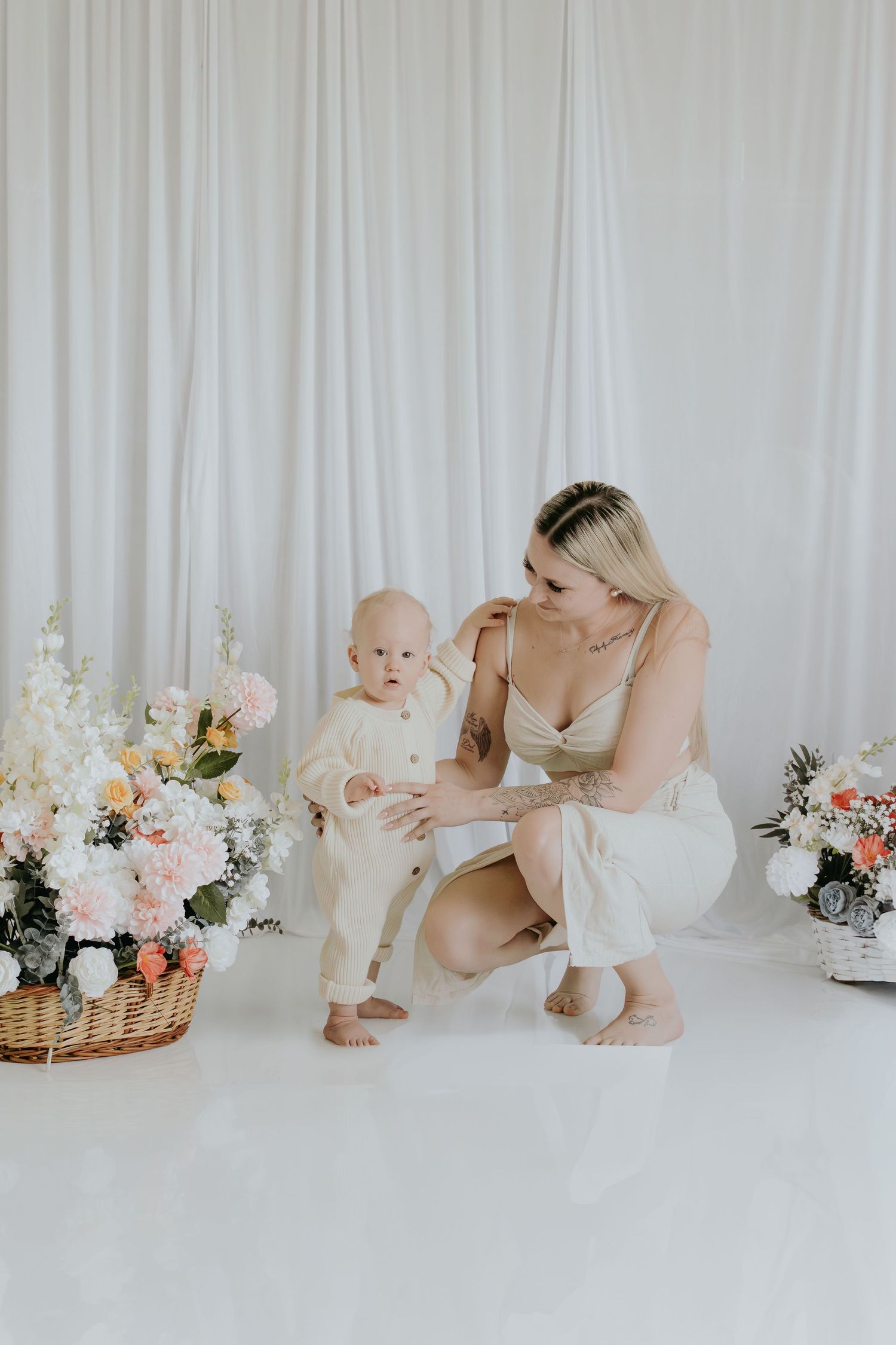A young mother crouches beside her baby near floral arrangements in a bright, white studio setting.