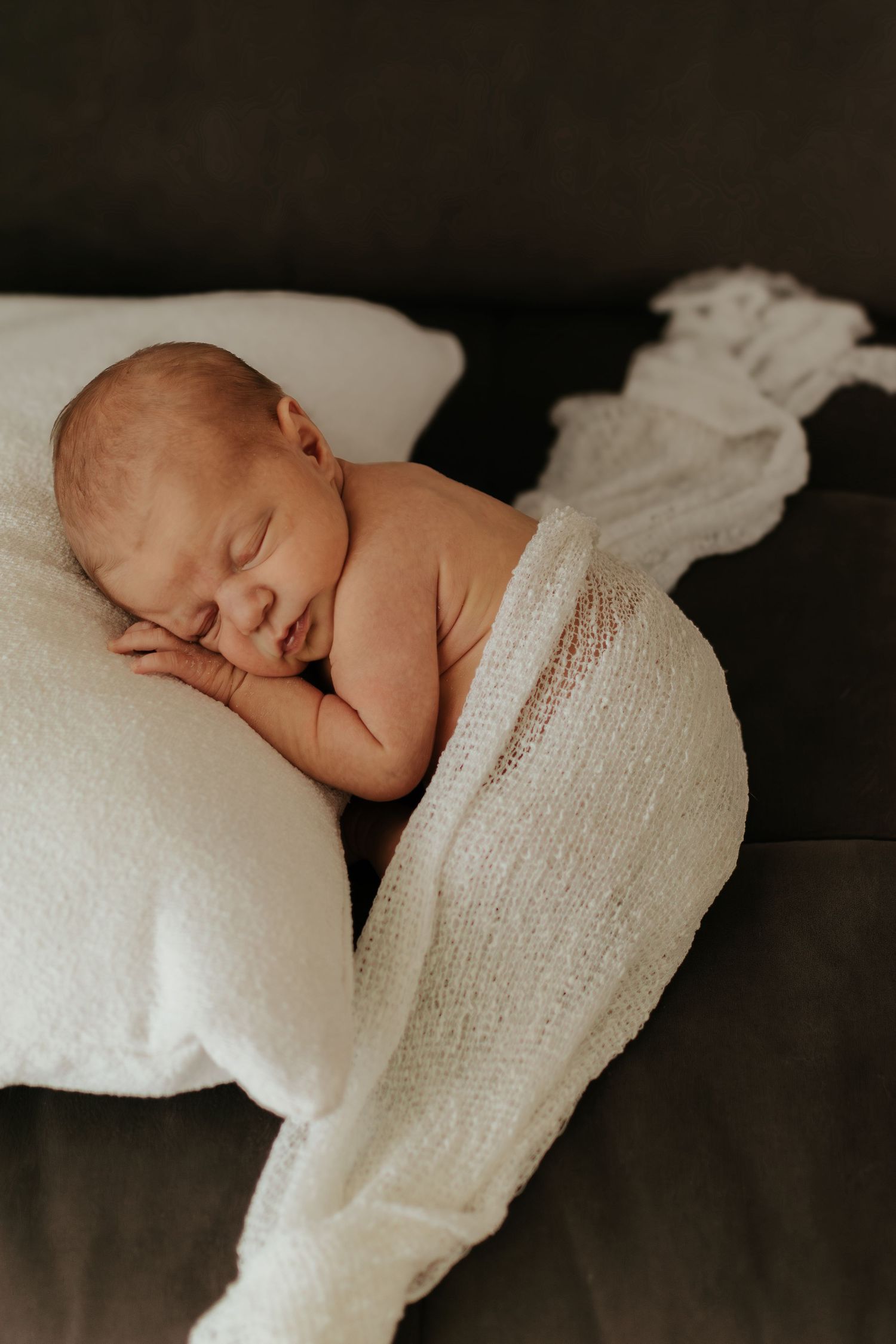 Peaceful newborn baby sleeping on a white pillow wrapped in a soft white knit blanket on a dark background.