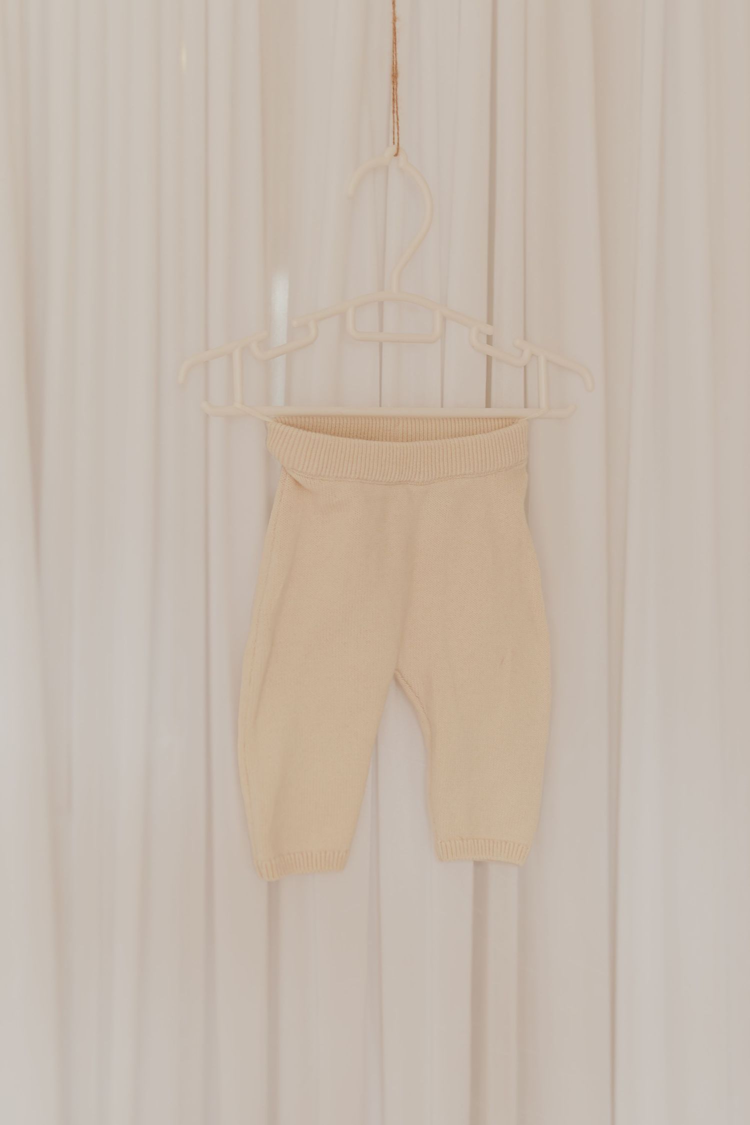 Eine beige Babyhose auf einem einfachen Drahtbügel vor einem hellen Vorhanghintergrund.