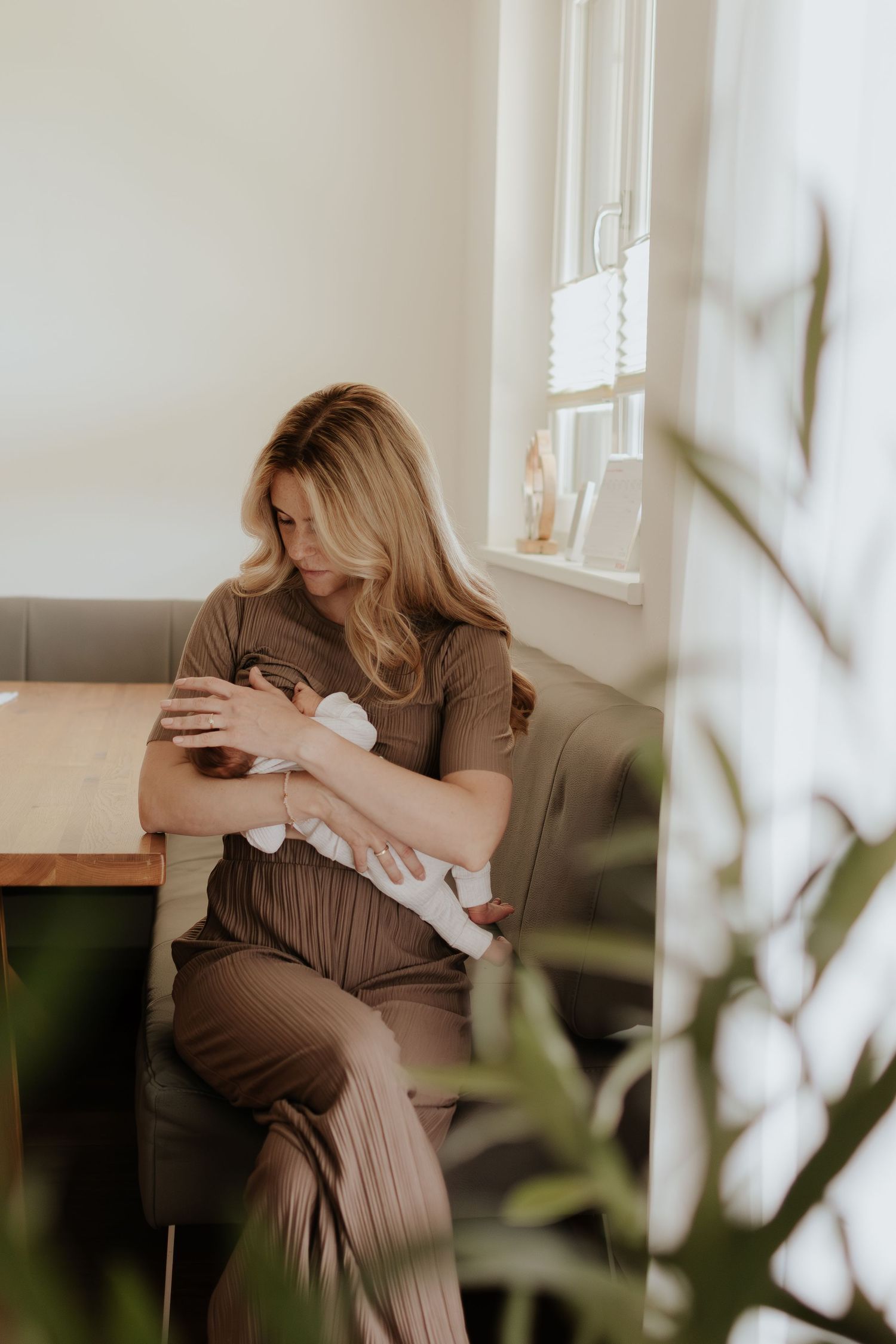 A mother with blonde hair breastfeeds her newborn baby while seated by a sunlit window in a cozy home setting.