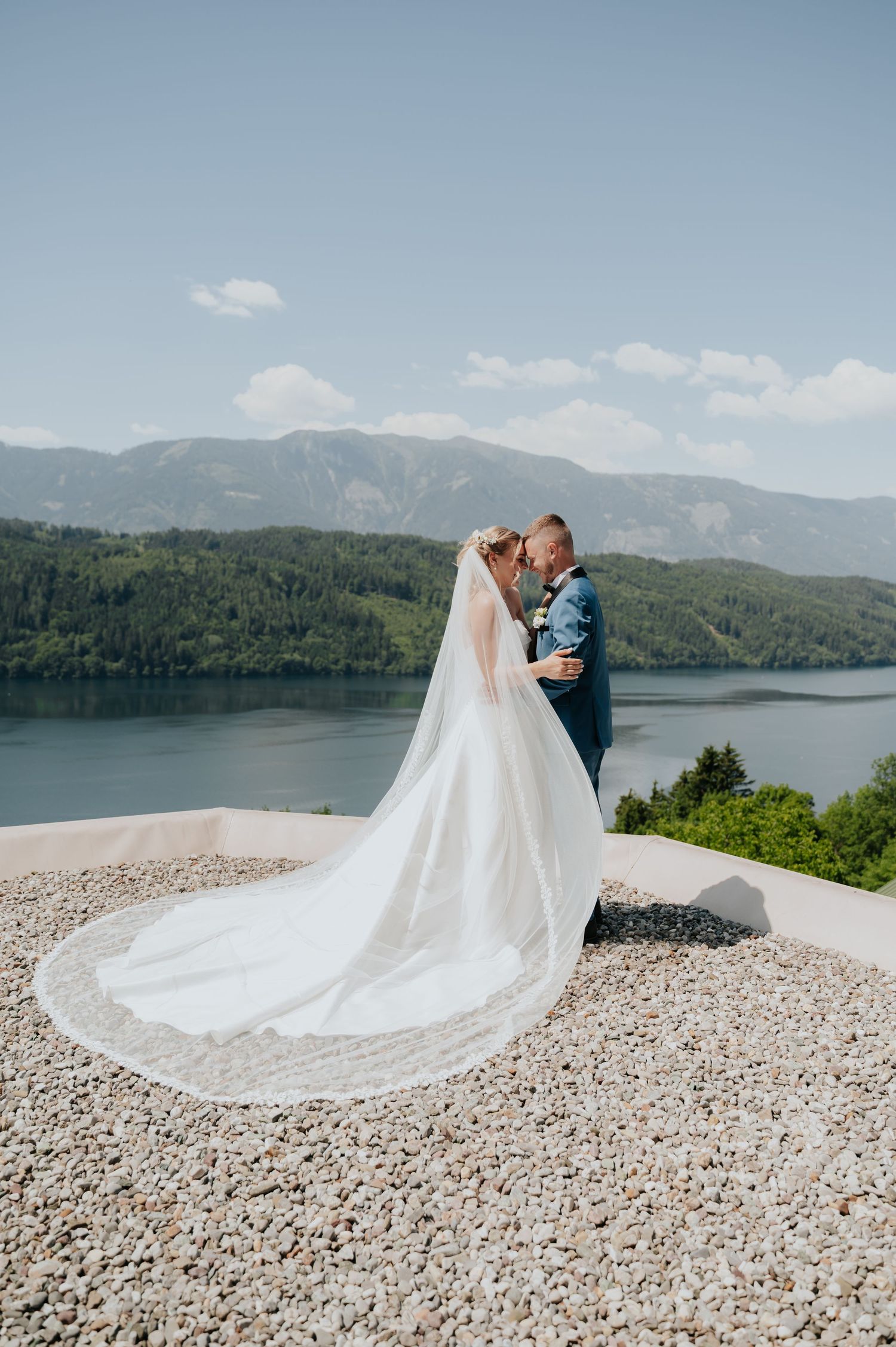 Bride and groom embrace on gravel terrace with stunning lake and mountain backdrop on their wedding day.