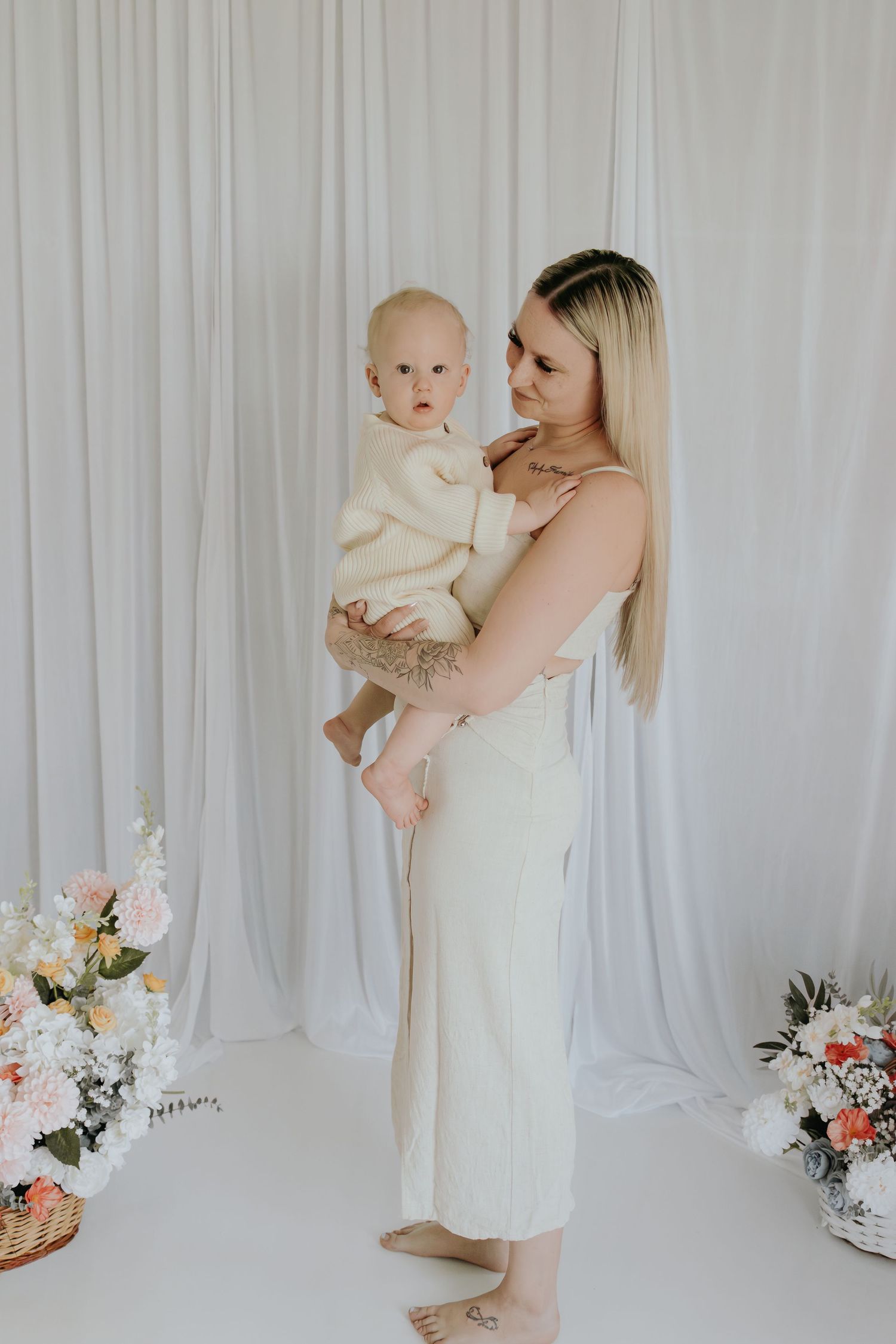 A blonde mother in a cream dress holds her baby in a white studio with floral arrangements.
