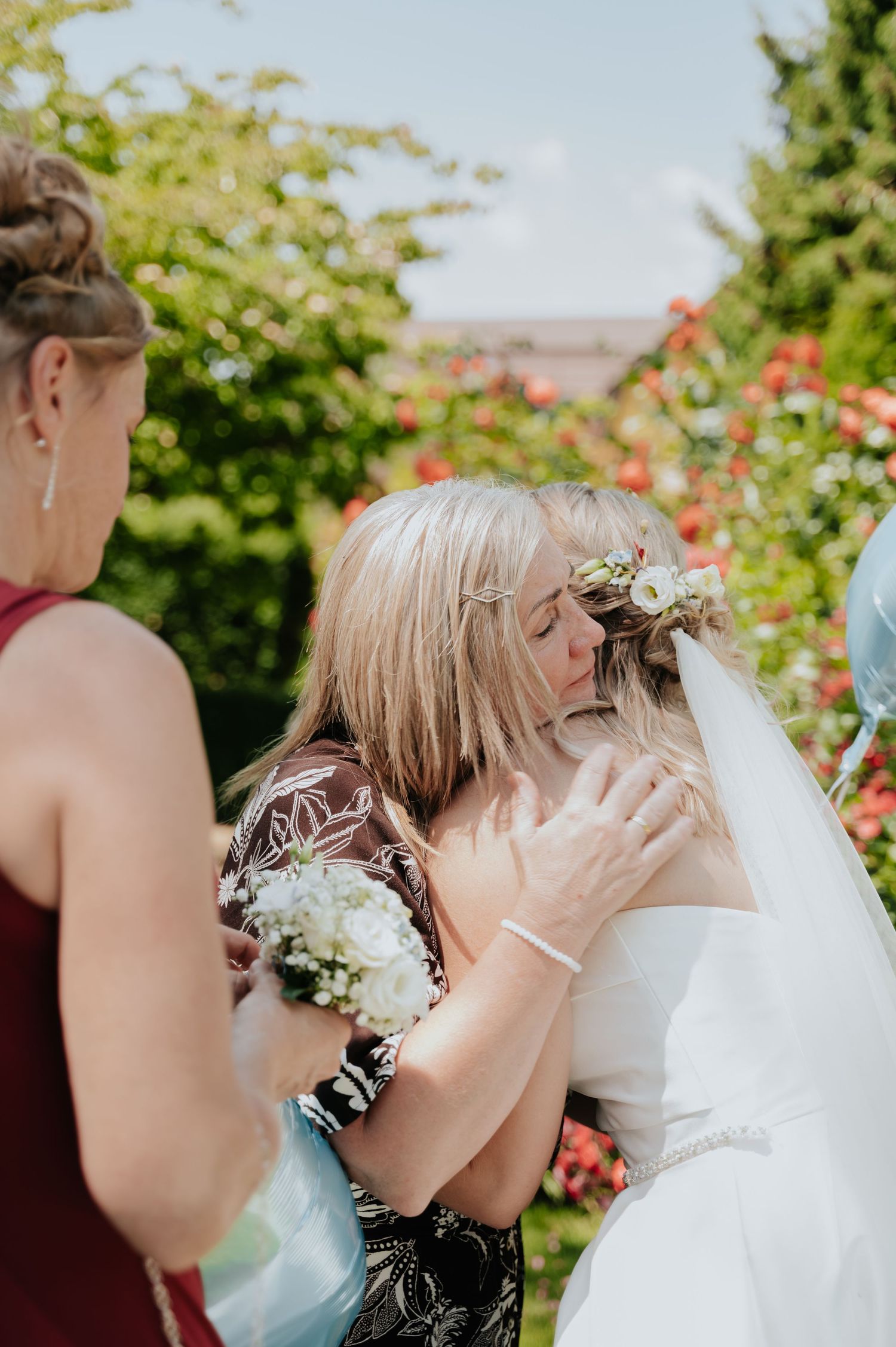 A bride in a white gown and floral veil embraces a blonde woman in a garden during an outdoor wedding ceremony.