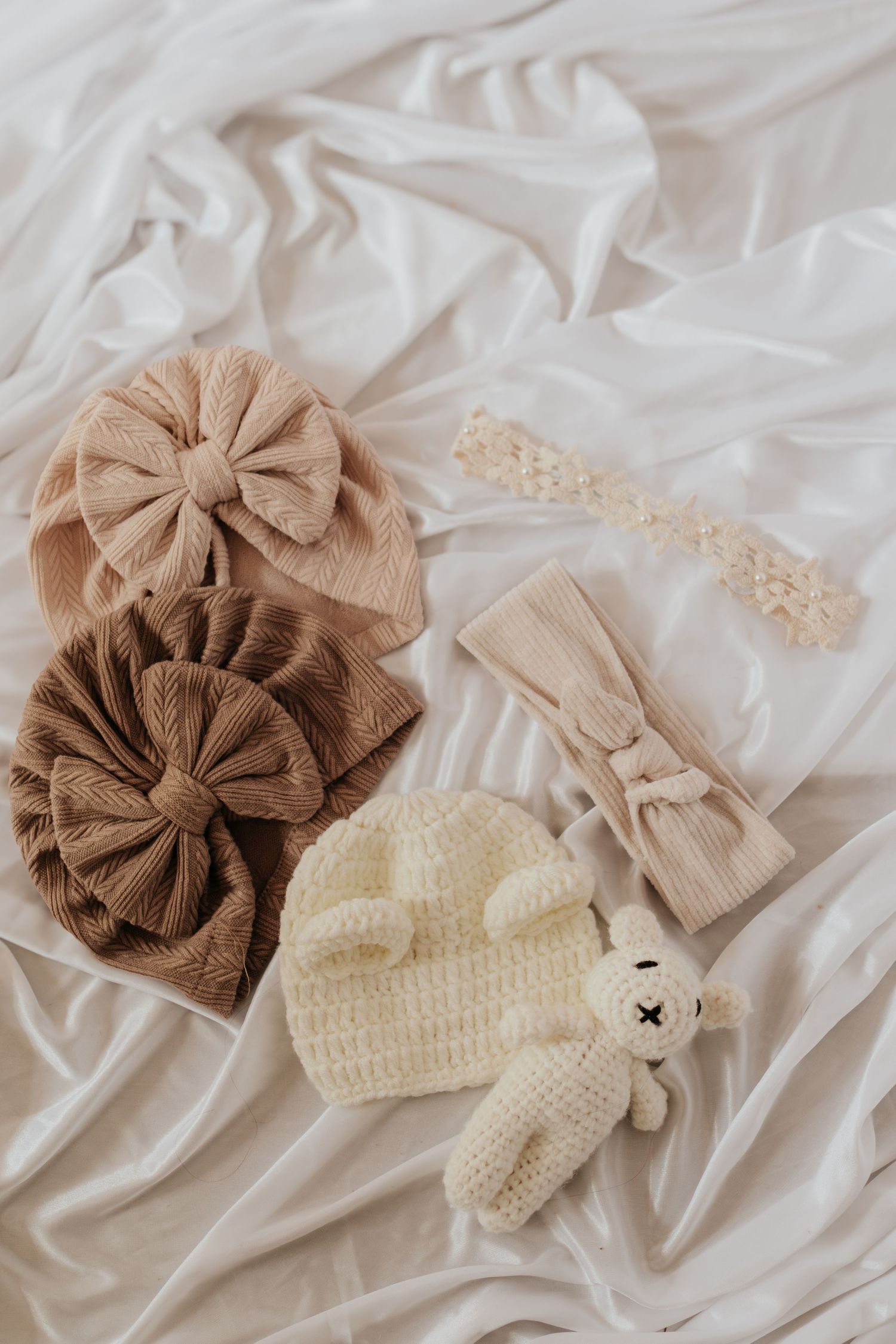 Neutrale Babyaccessoires, darunter Strickmützen mit Schleifen, Stirnbänder und ein kleines Plüschtier auf weißem Stoff.