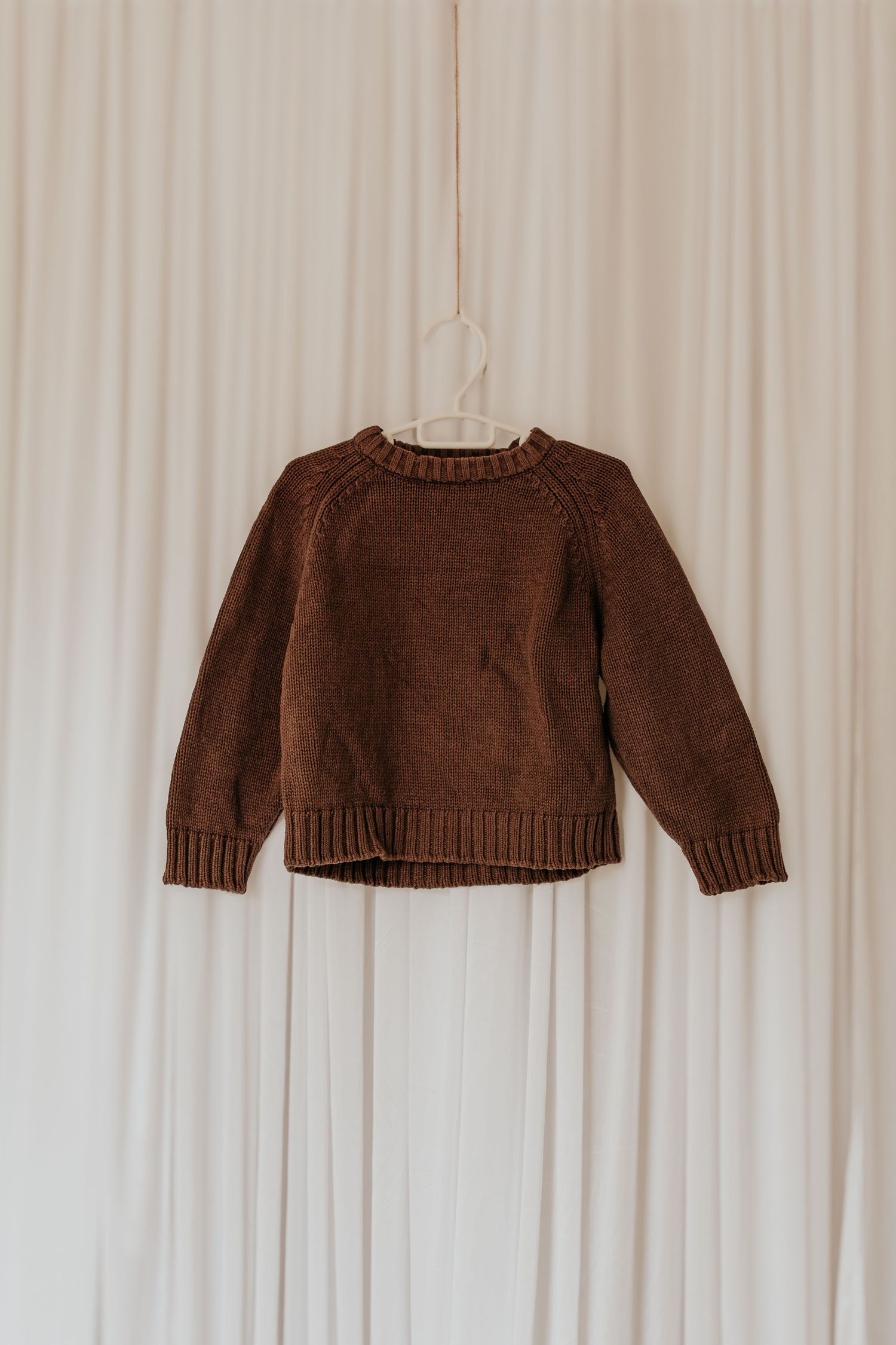 Ein brauner Strickpullover, der vor einem weißen Vorhanghintergrund hängt.