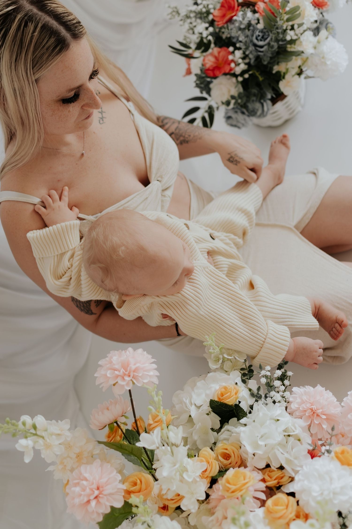 Tattooed blonde mother in white breastfeeding her baby surrounded by colorful floral arrangements in a bright studio setting.