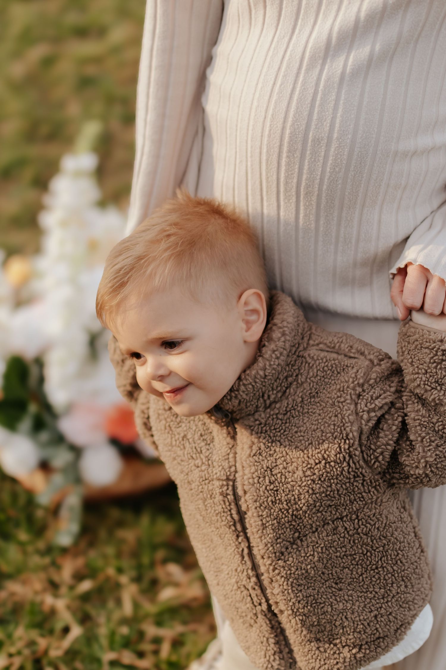 A smiling toddler in a brown teddy bear jacket holds onto their mother's hand outdoors near flowers.