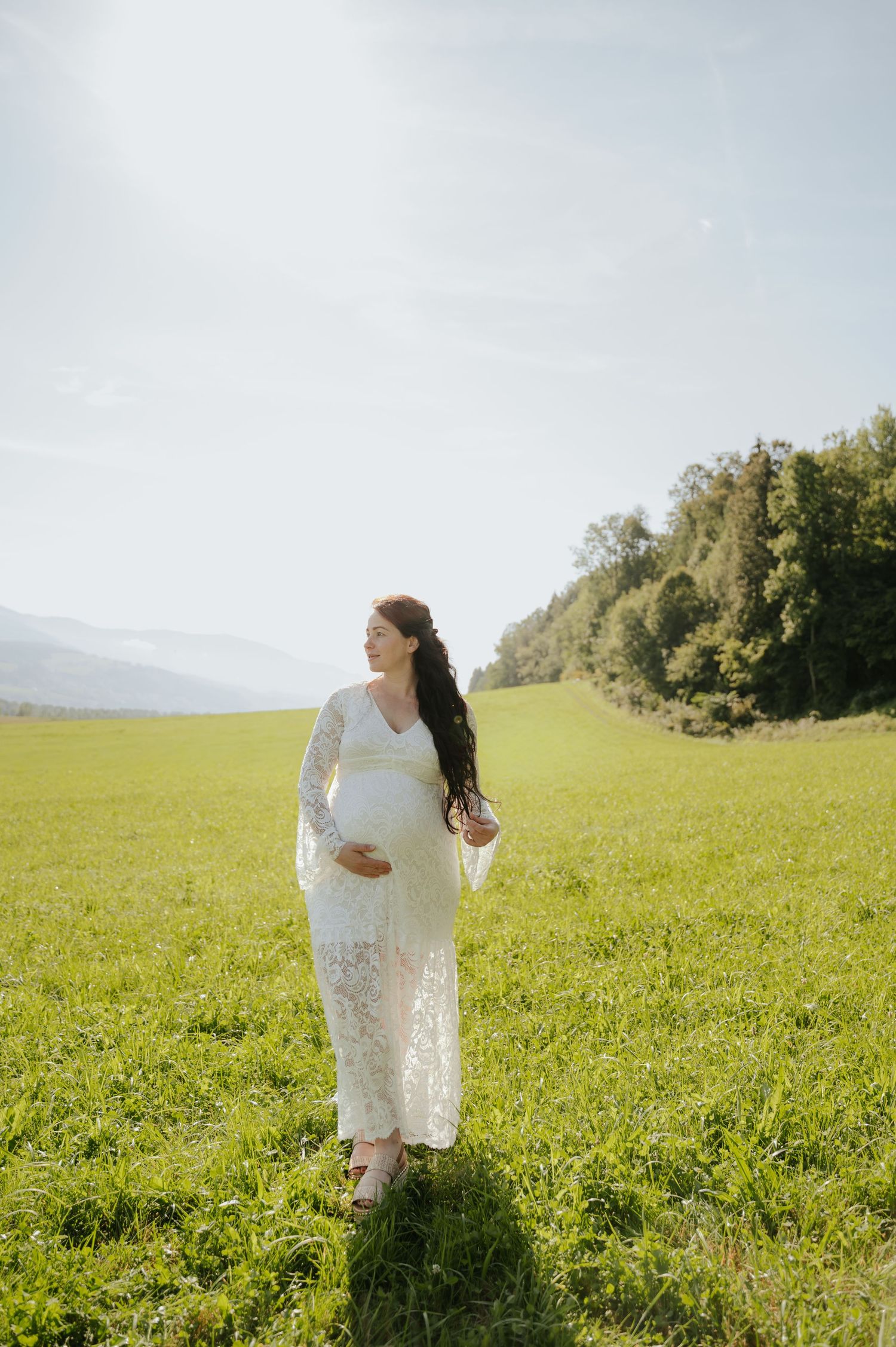 Pregnant woman in white lace dress standing in green meadow with mountains and trees in background.