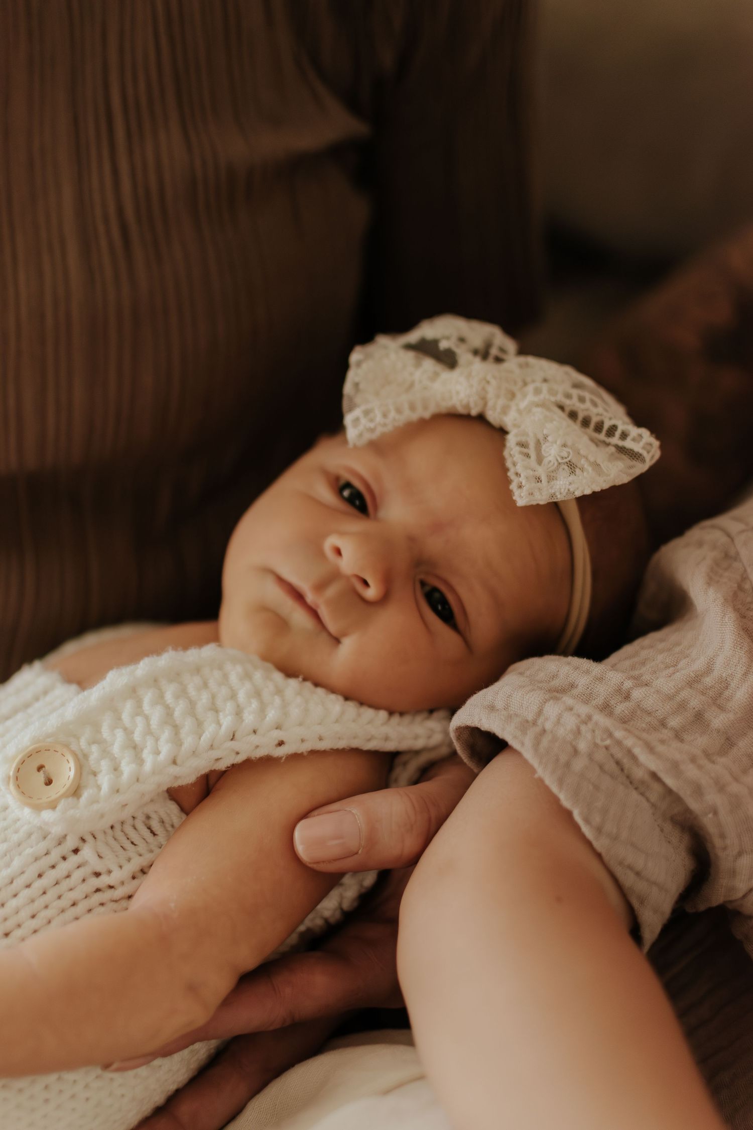 Newborn baby girl in white knit romper and lace bow headband resting in mother's arms on brown sofa.