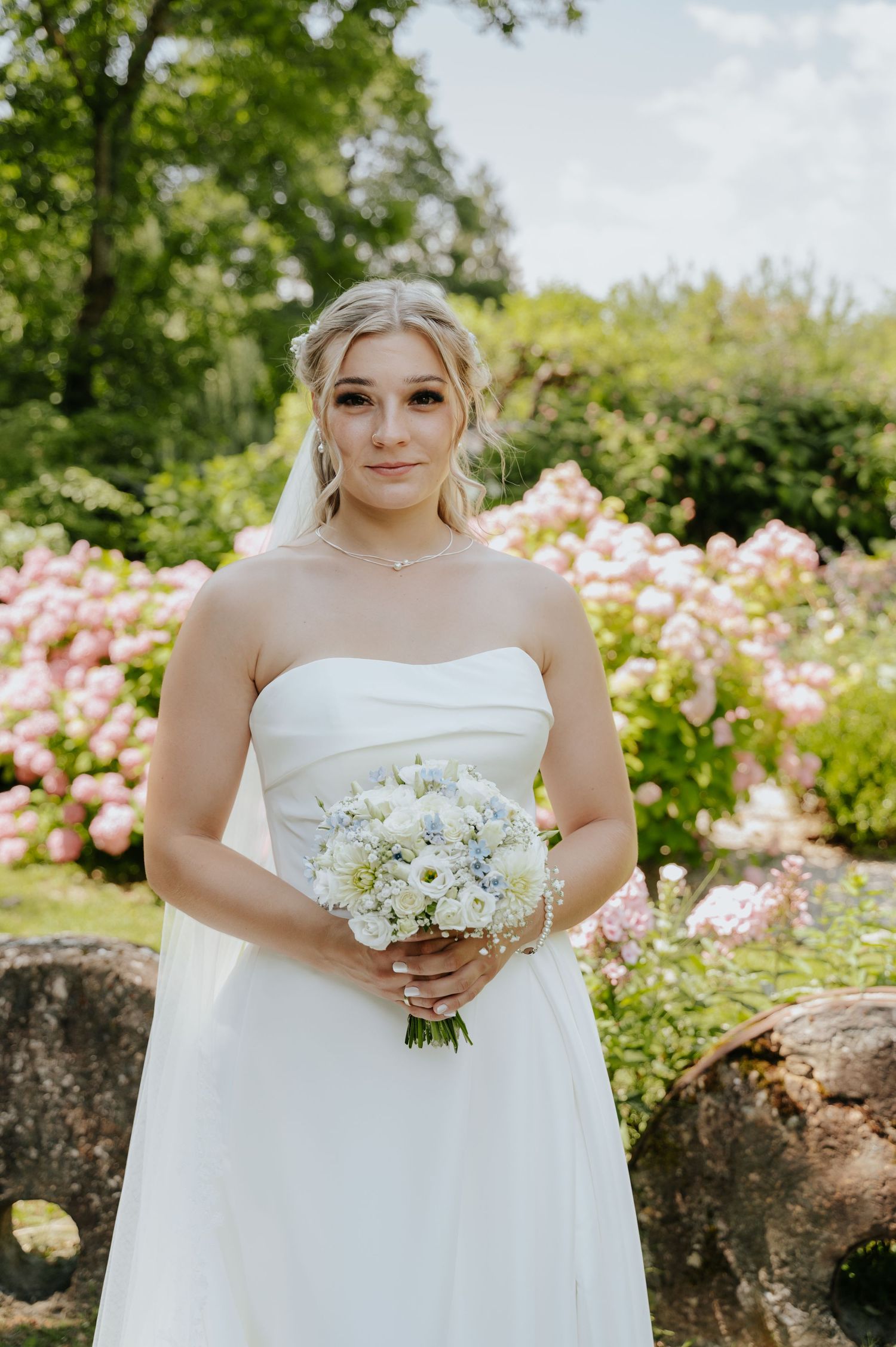 Elegant bride in strapless white gown holds blue and white bouquet before blooming pink hydrangeas.