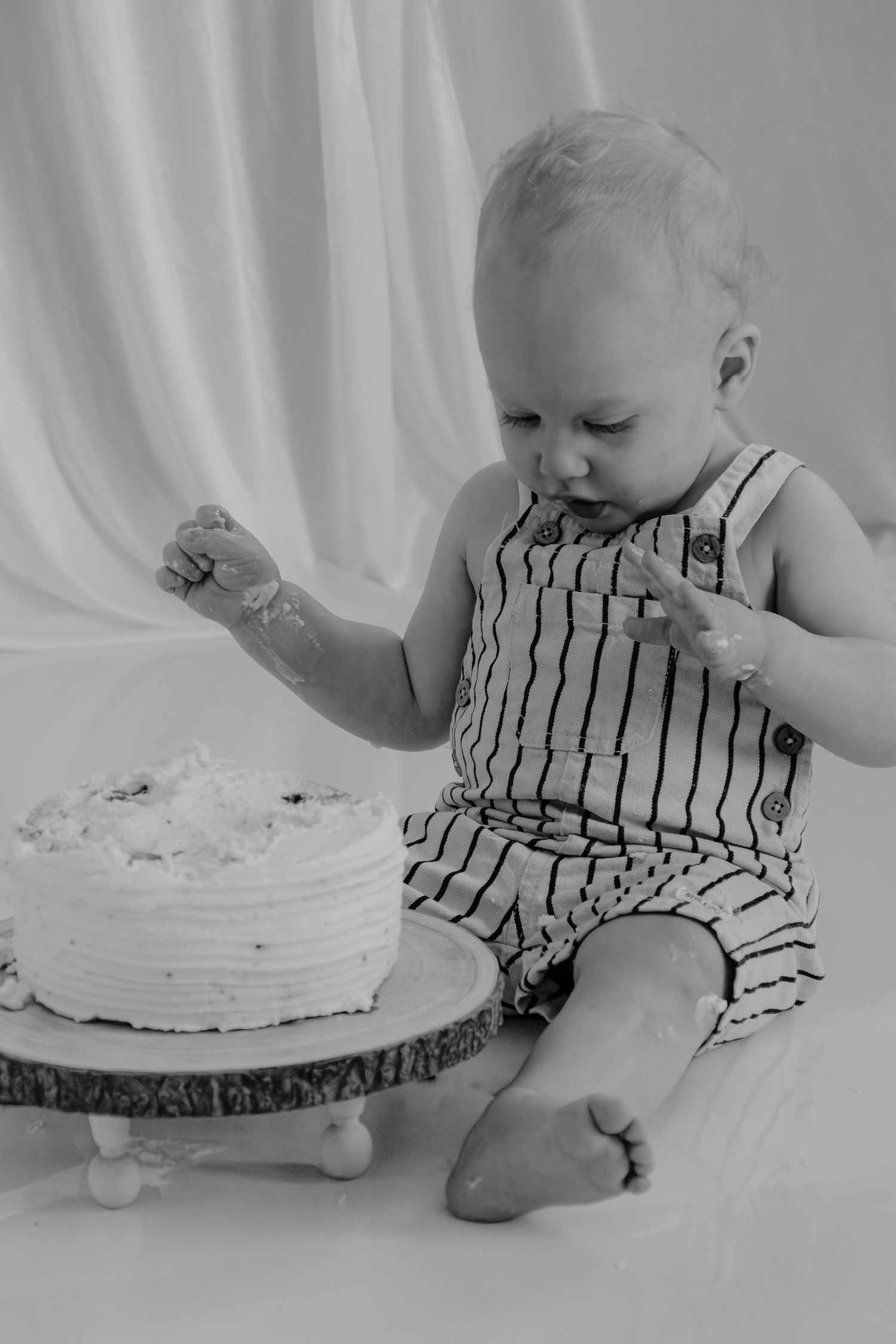 Black and white photo of a baby boy in striped overalls smashing his first birthday cake on a wooden cake stand.