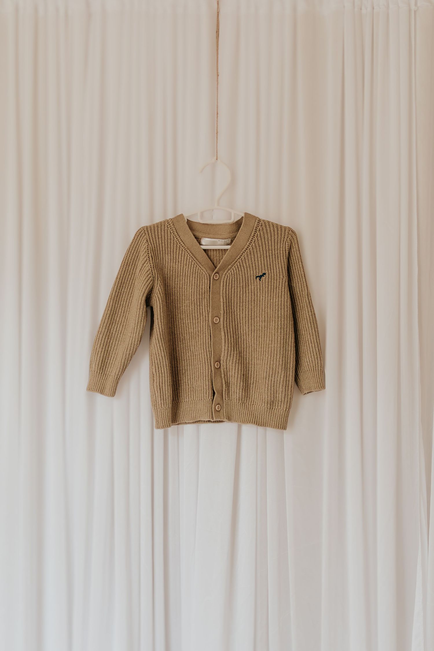 Hellbrauner gerippter Strickpullover mit Knopfverschluss vor weißem Vorhanghintergrund.