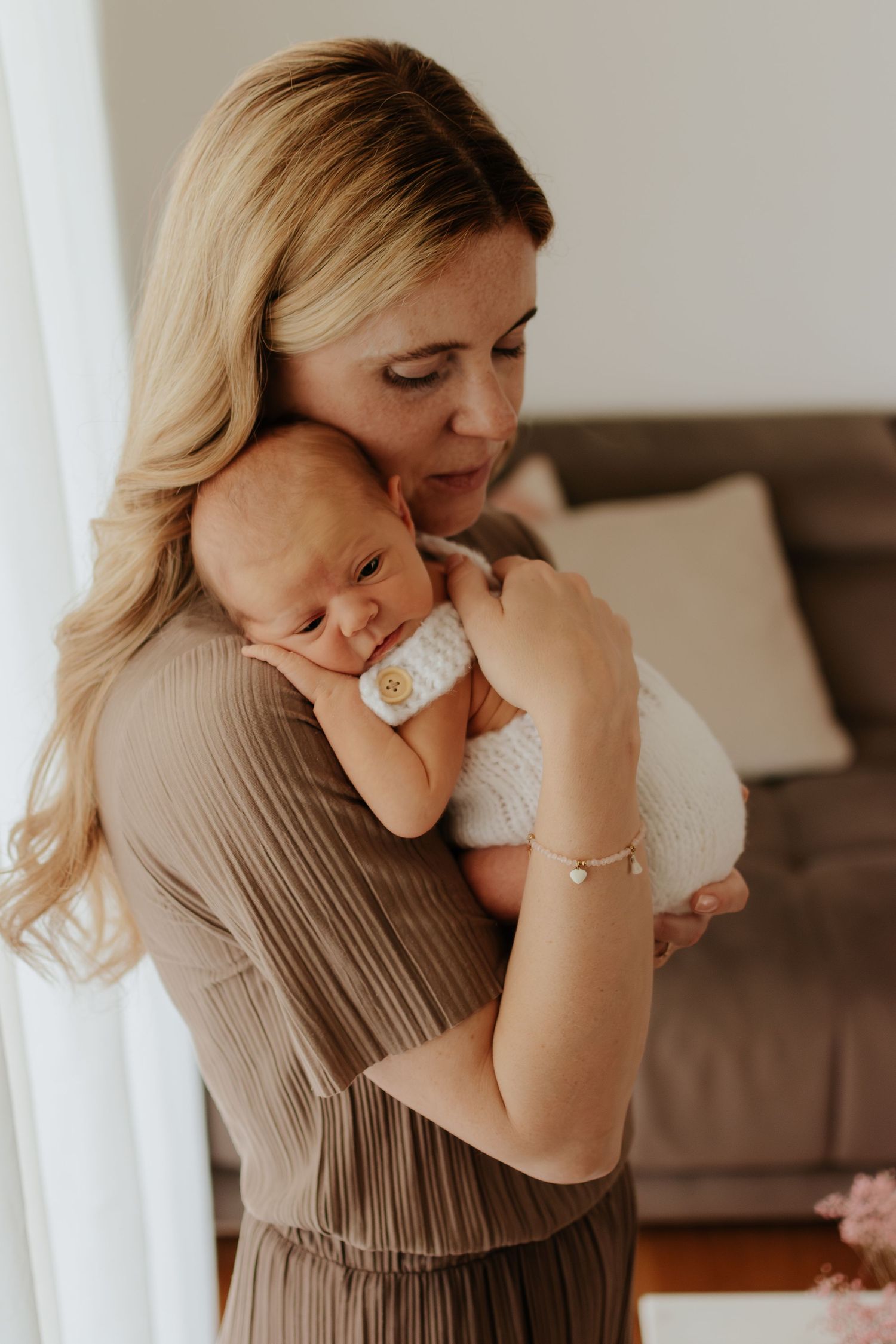 A blonde mother tenderly holds her sleeping newborn baby wrapped in white, standing in a cozy living room.