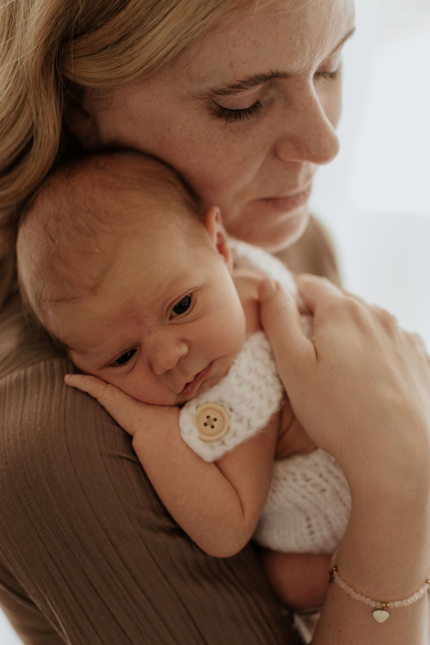 A tender moment as a mother cradles her newborn baby dressed in a white knit outfit against her shoulder.