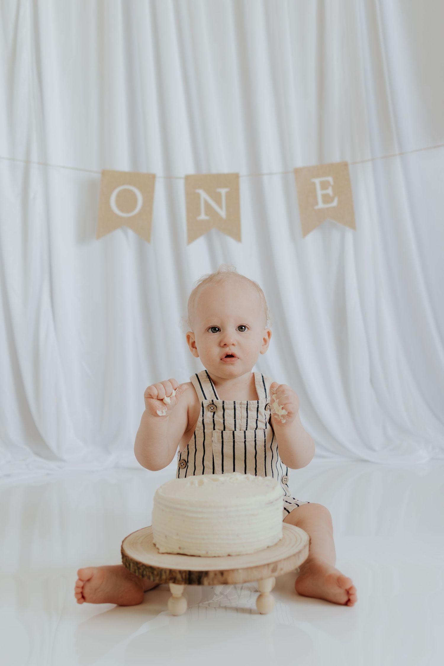 Baby's first birthday cake smash photo with white frosted cake, striped romper, and burlap ONE banner backdrop.