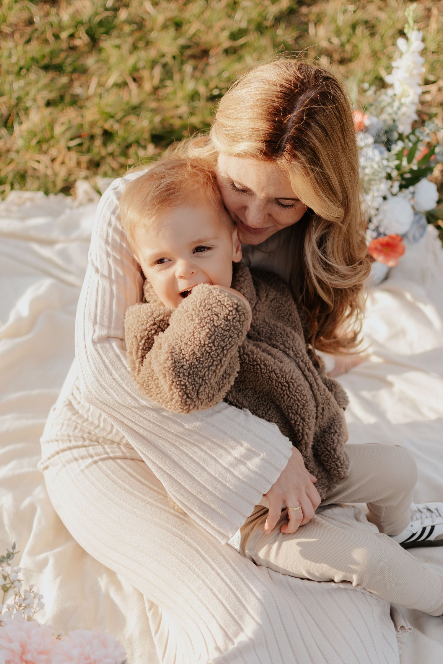 A mother hugging her red-haired toddler on a white blanket outdoors in warm sunlight.
