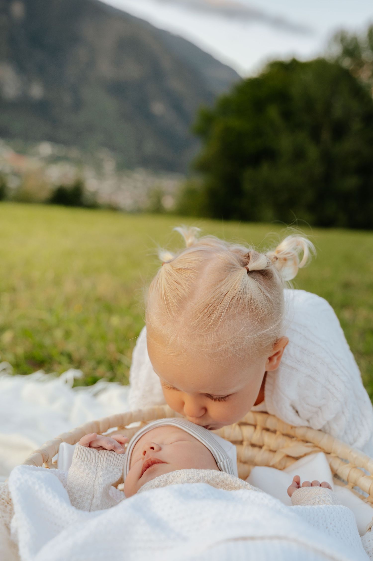 Toddler girl with blonde hair gently kissing sleeping newborn baby in wicker basket on white blanket outdoors.