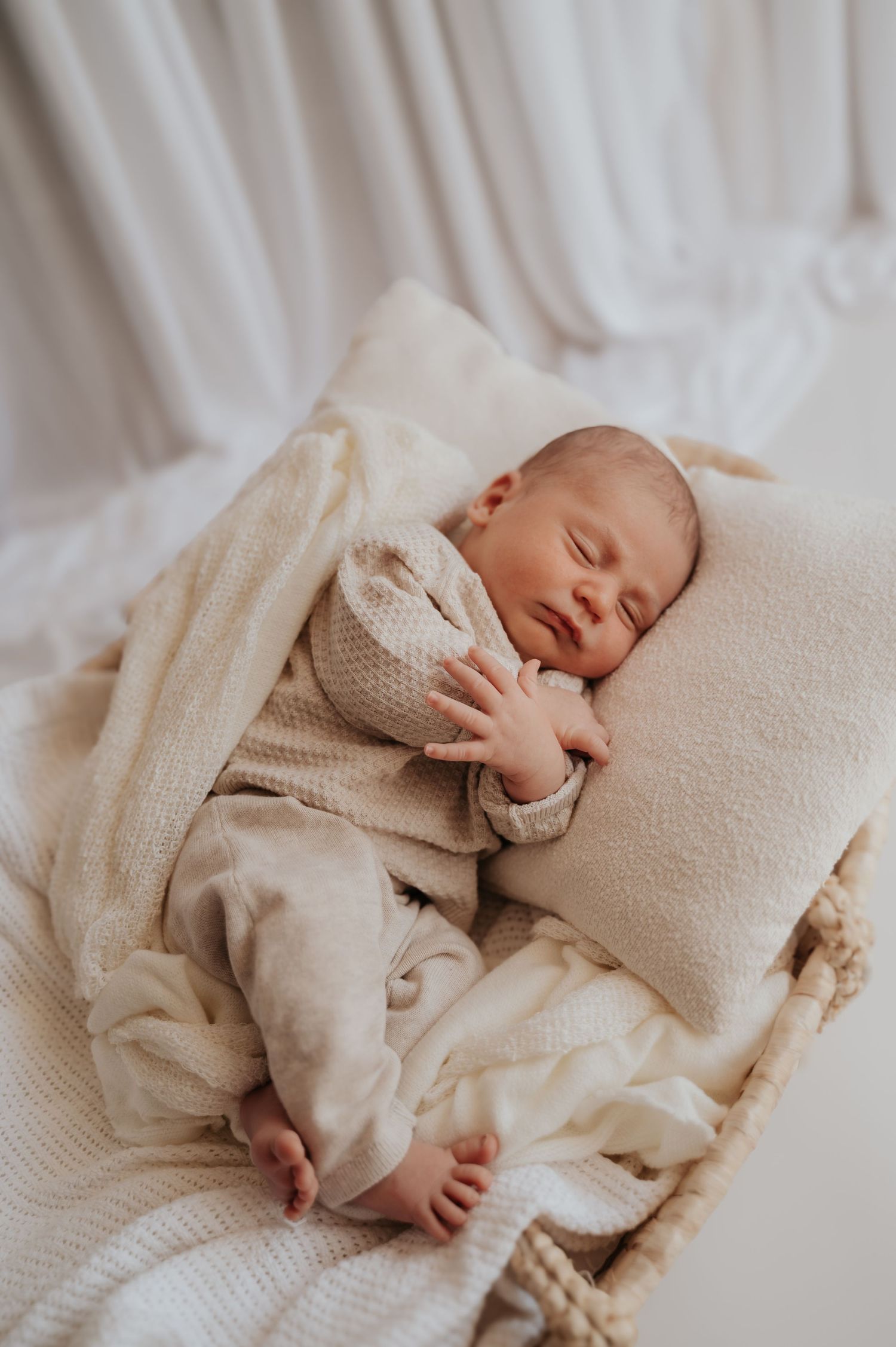 A peaceful newborn baby sleeping in a wicker bassinet on cream blankets and pillow in soft white light.