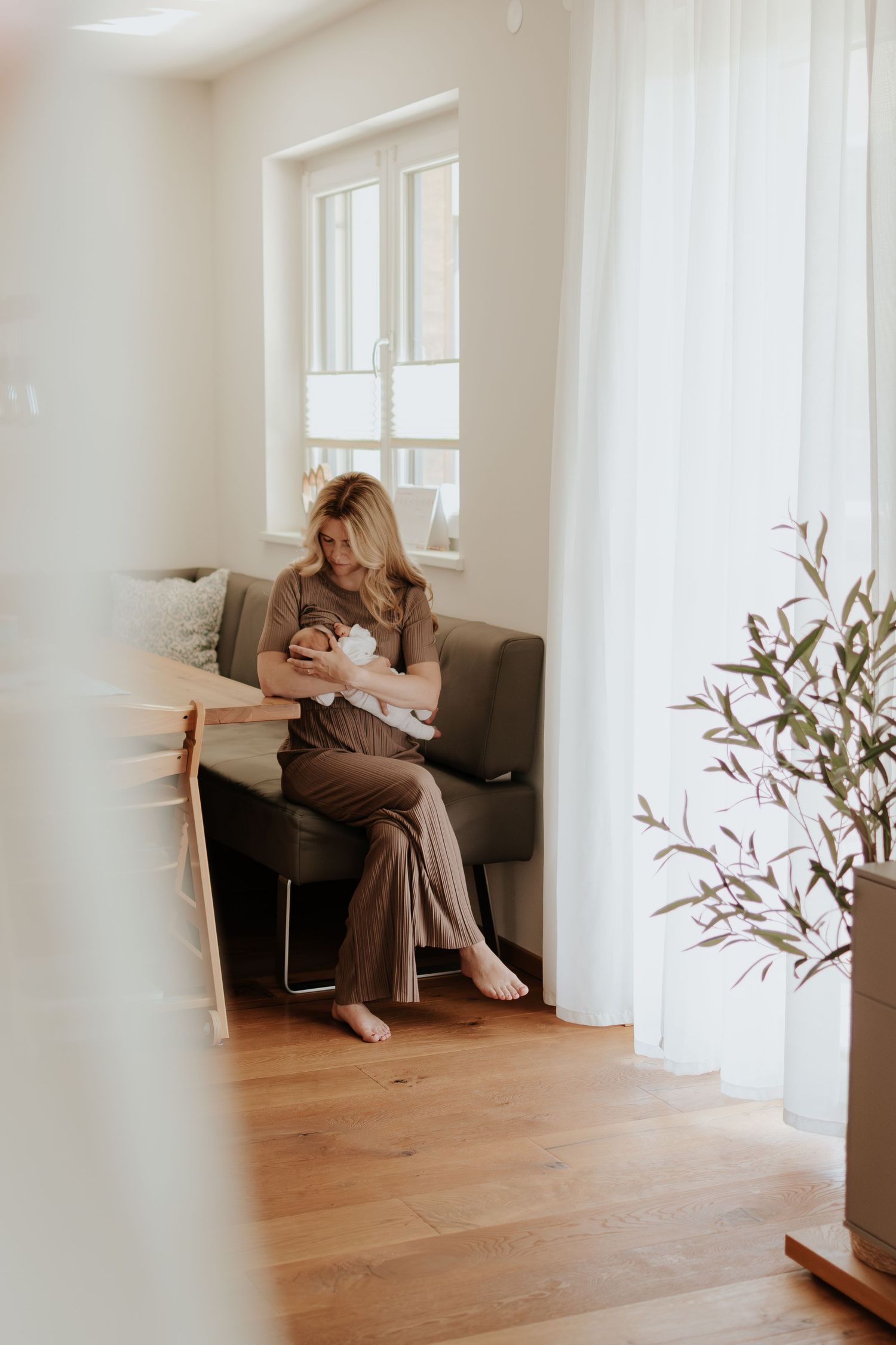 A woman in brown outfit sits on a gray sofa, cradling a newborn baby in a bright, modern living room.