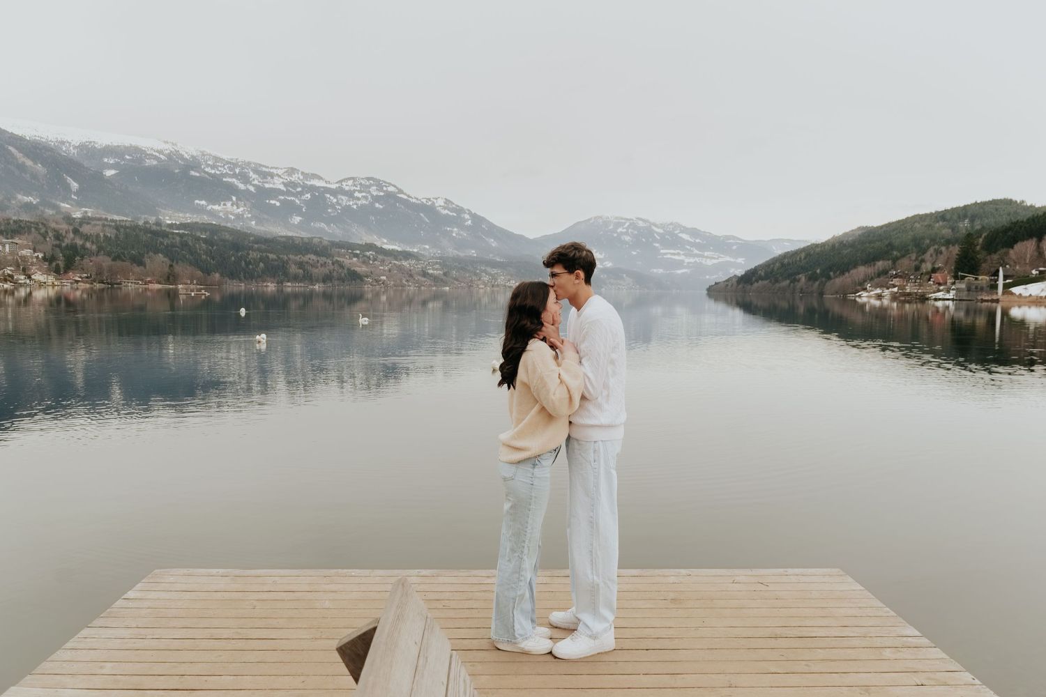 Couple kissing on a wooden dock with a serene mountain lake and snow-capped hills in the background.