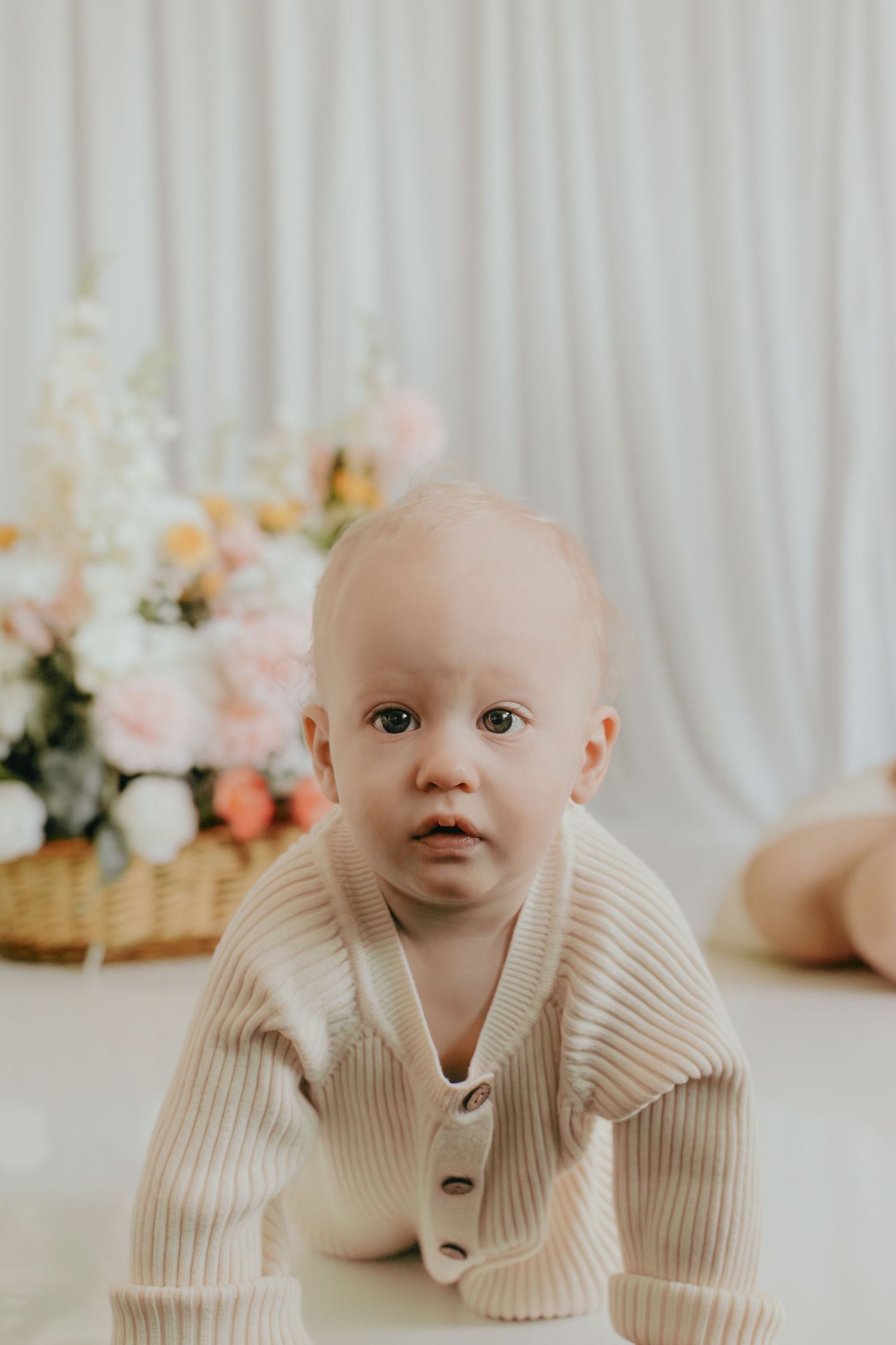 Adorable baby in a beige ribbed knit cardigan crawling on the floor with soft floral arrangement in background.