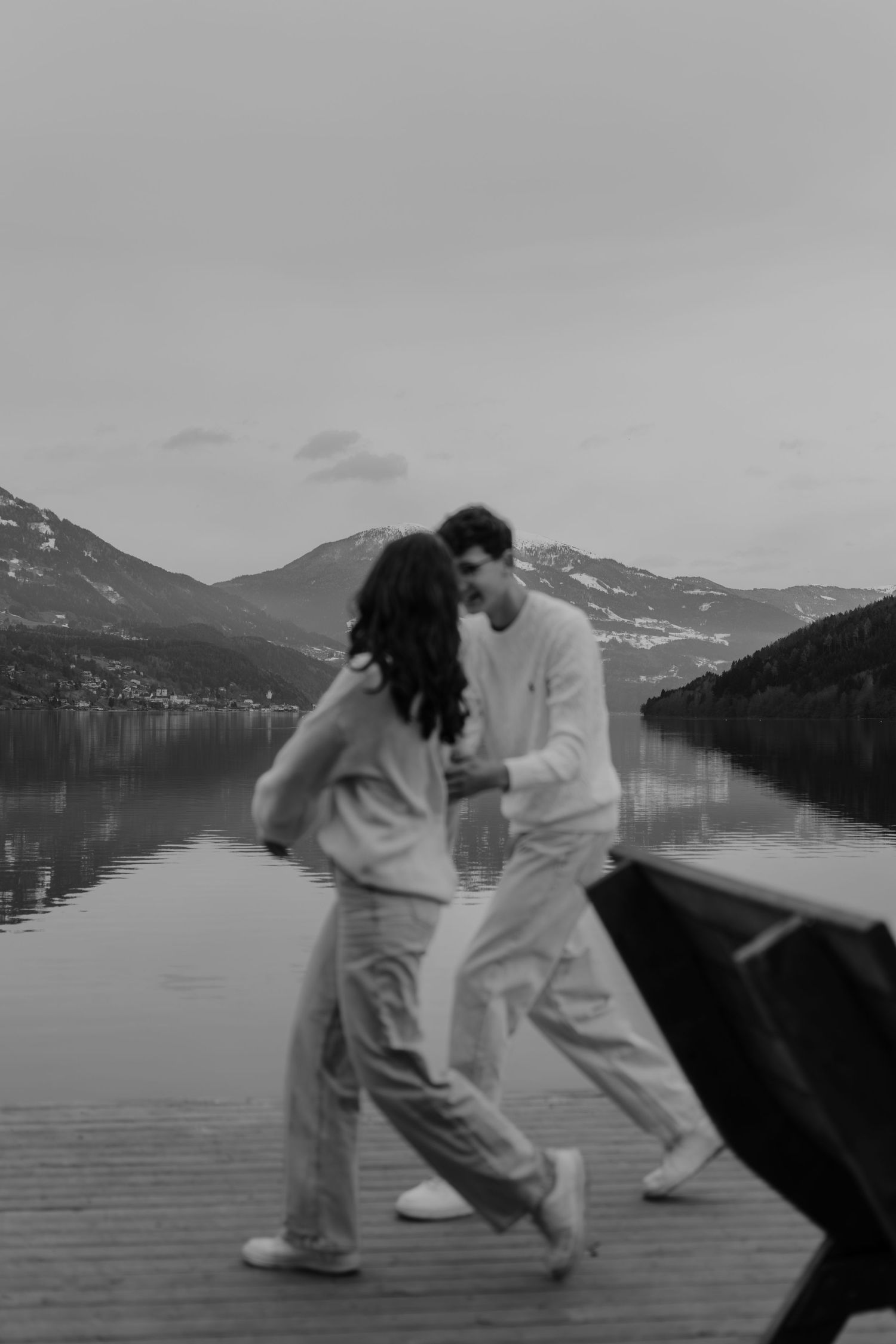 Ein verspieltes Paar in Schwarz-Weiß-Foto zusammen auf einem Dock mit Bergen im Hintergrund.