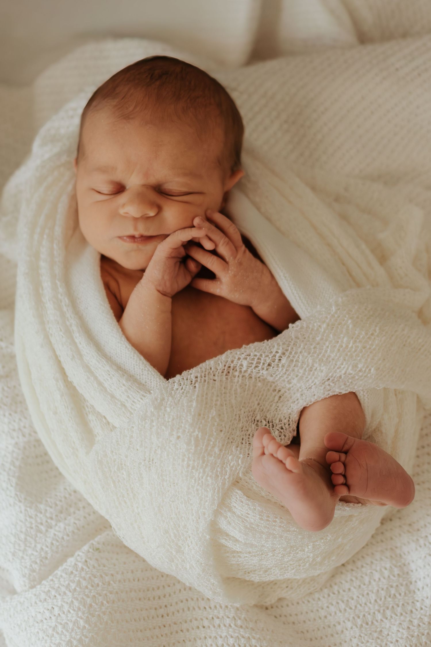Peaceful newborn baby sleeping wrapped in a soft white knit blanket with tiny hands and feet visible.
