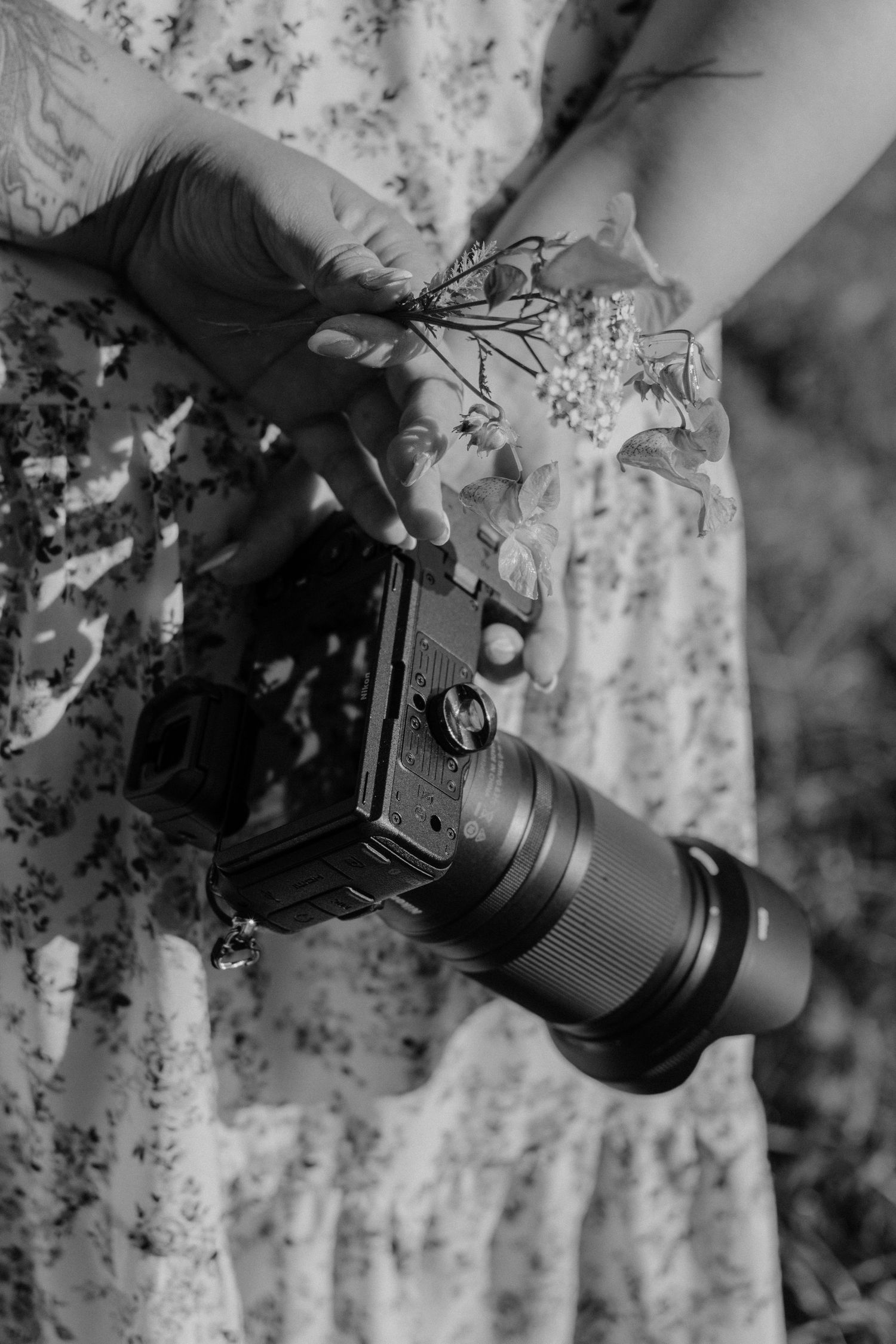 Black and white photo of a woman in a floral dress holding a camera with a telephoto lens and small wildflowers.