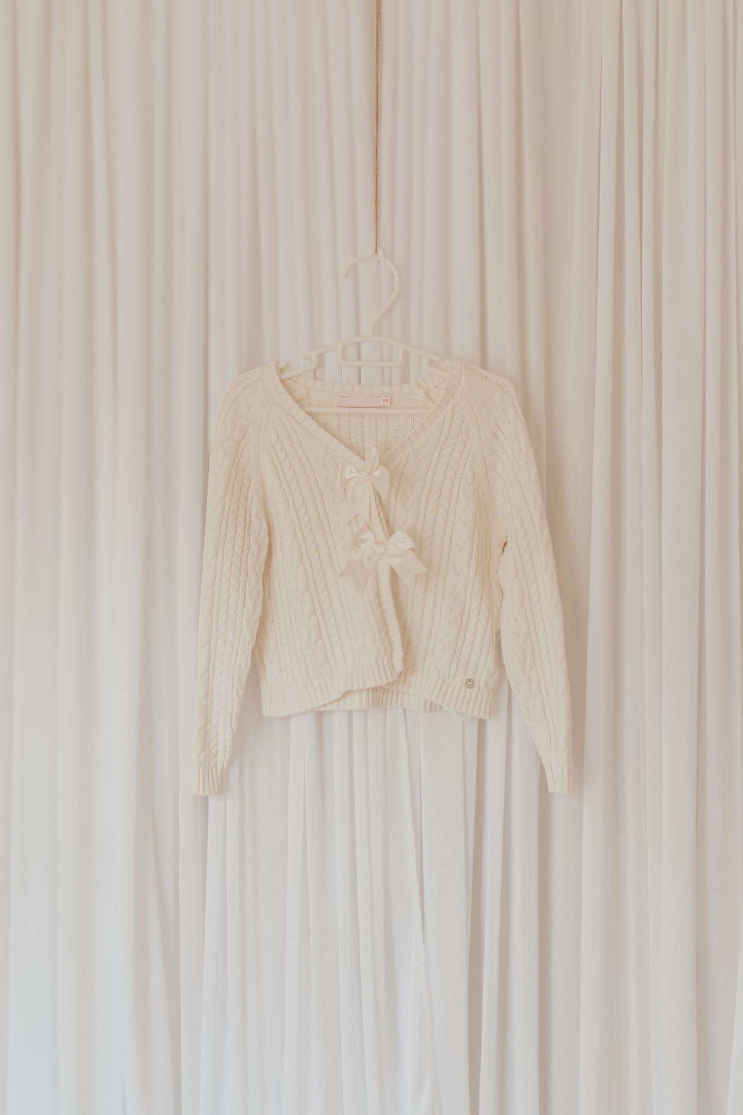 Ein cremefarbener Strickpullover mit Krawattenverschluss, der an einem weißen Plastikaufhänger vor einem hellen Vorhanghintergrund hängt.