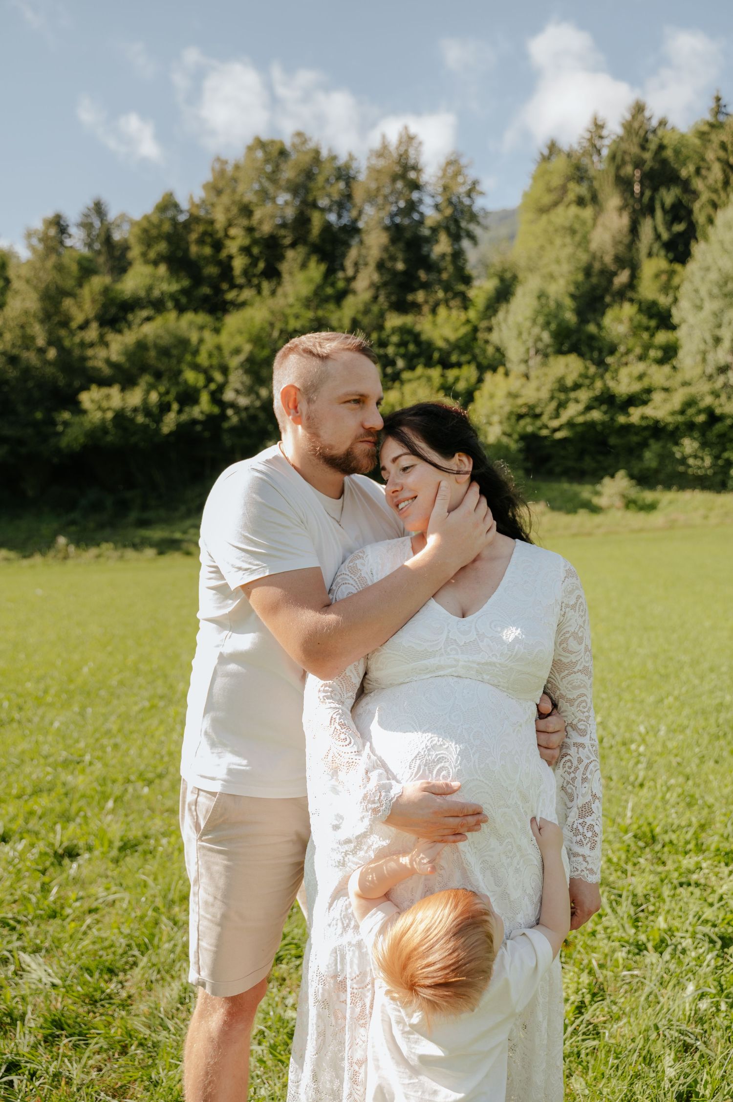 Pregnant couple embracing in a green meadow, man in white shirt kissing woman in lace dress, sunny day.